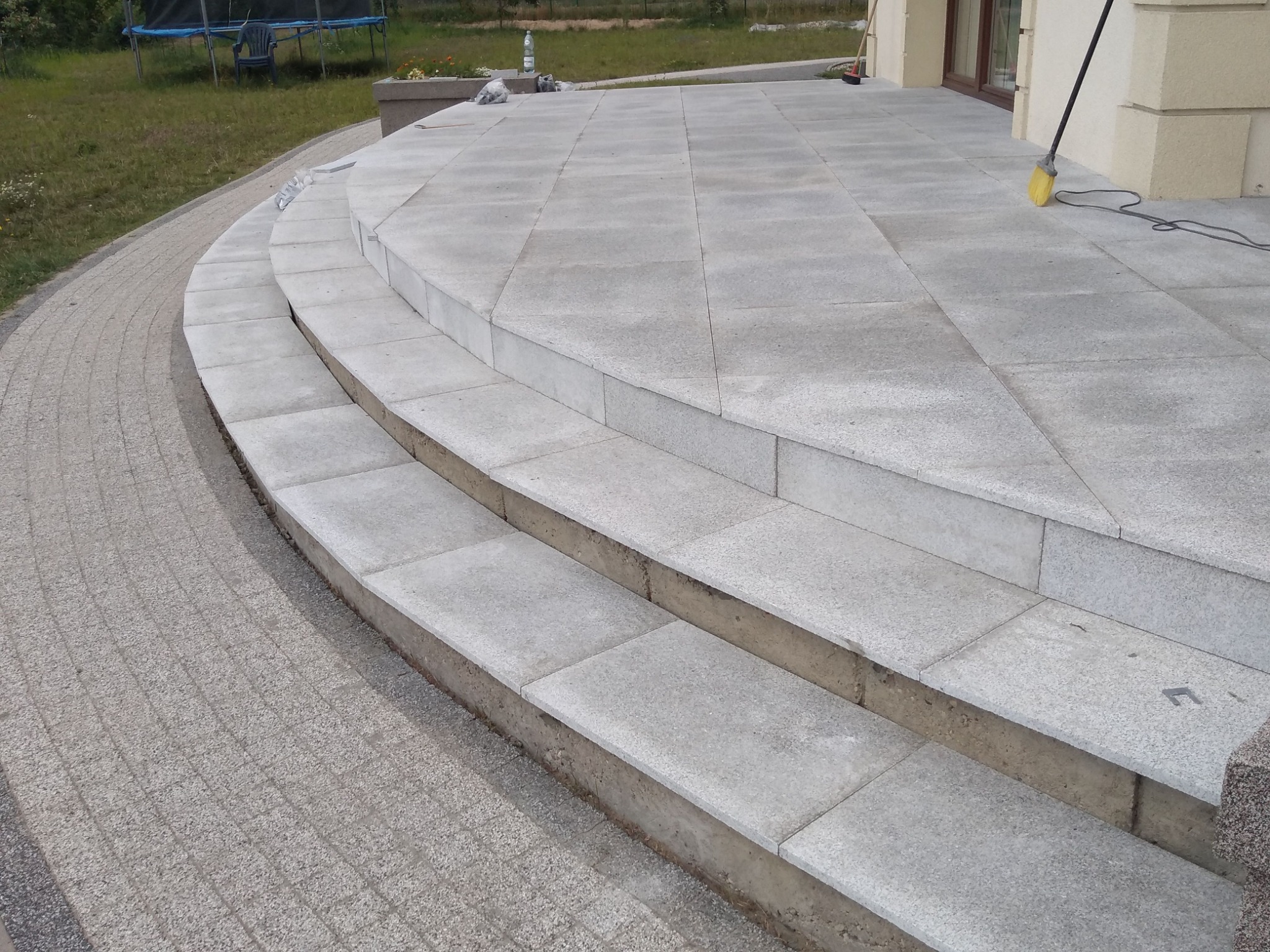 Wykończenie terenu przy domu: Schody i taras wyłożone szarą kostką granitową, widoczny fragment chodnika z kostki brukowej oraz budynek z oknem i elementami elewacji. Na tarasie stoi miotła.