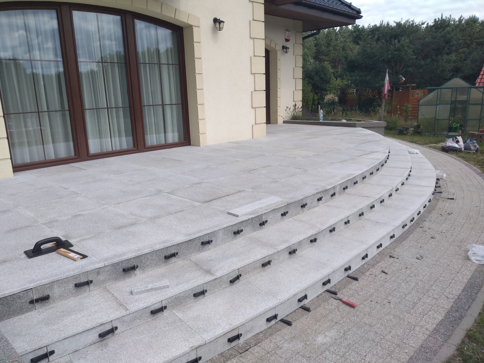 Granitowe schody zewnętrzne w trakcie montażu, widoczne czarne elementy dystansowe i narzędzia glazurnicze, dom z kremową elewacją w tle.