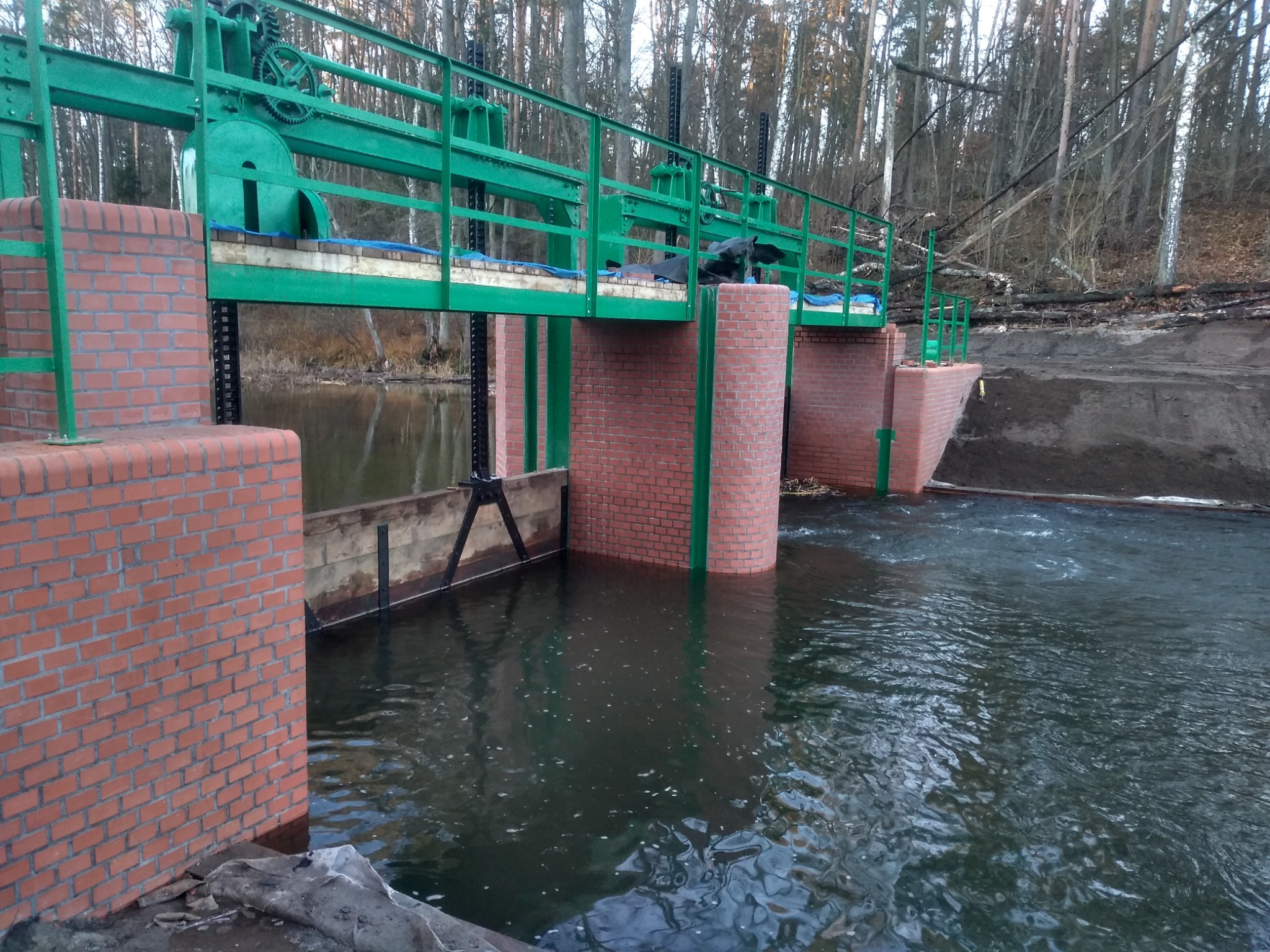 Brązowo-czerwona konstrukcja z cegły i zielonych metalowych elementów nad przepływającą wodą, w tle drzewa.