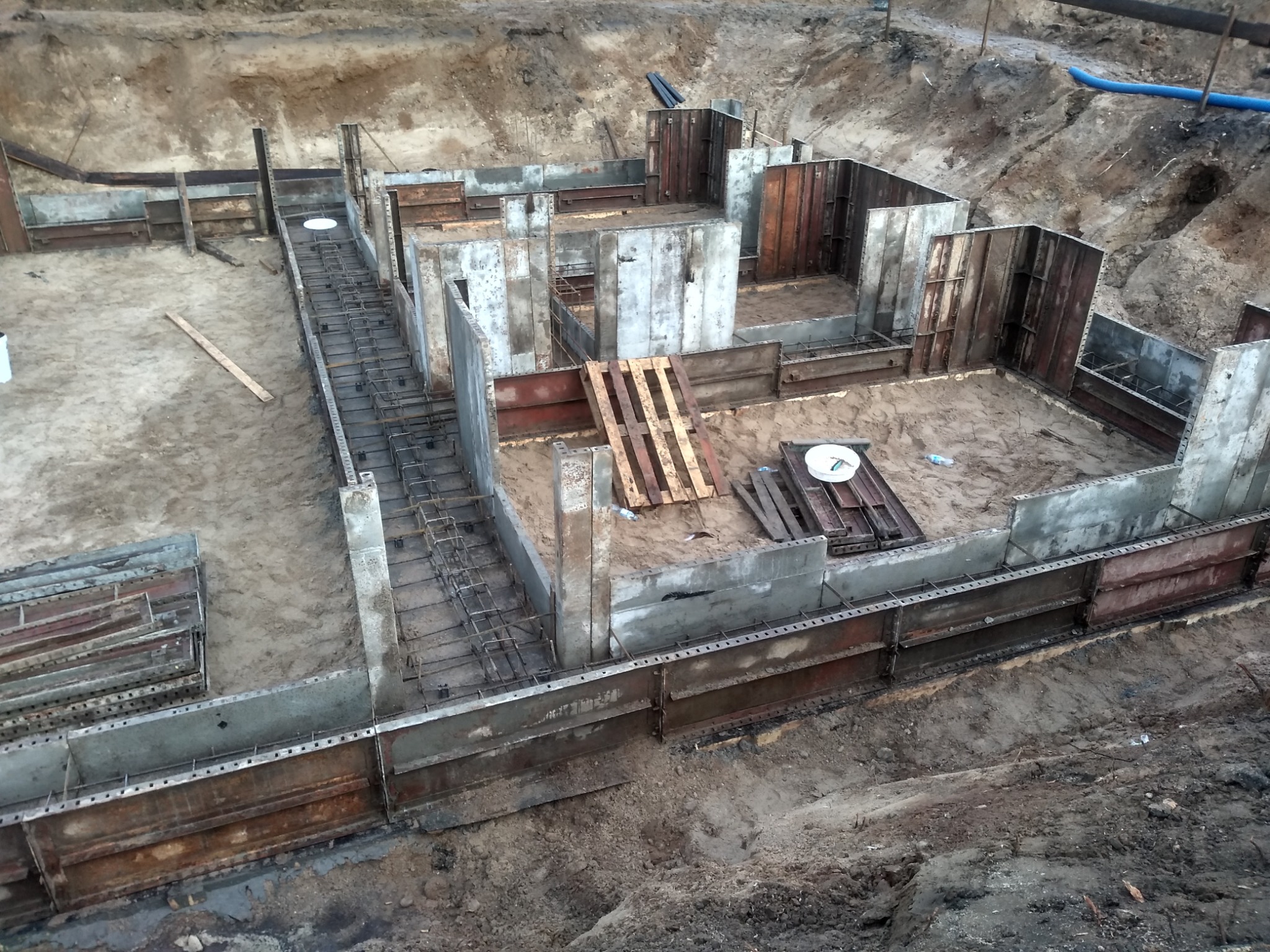 Zalane betonem fundamenty domu w trakcie budowy, widoczne szalunki i zbrojenia na piaszczystym podłożu, z paletą i plastikową butelką w tle.