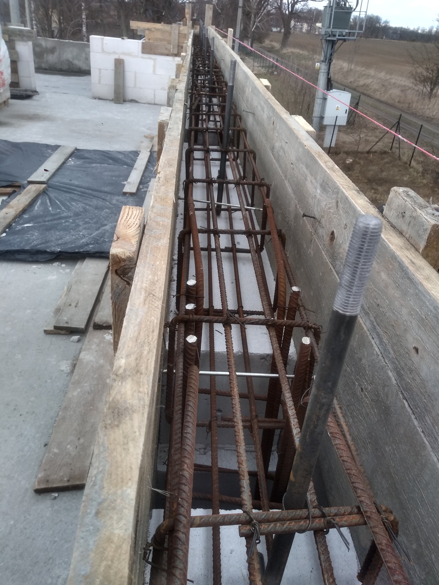 Zbrojenie w szalunku drewnianym przygotowane do zalania betonem, widok z góry na placu budowy, w tle bloczki betonowe i słup elektryczny.
