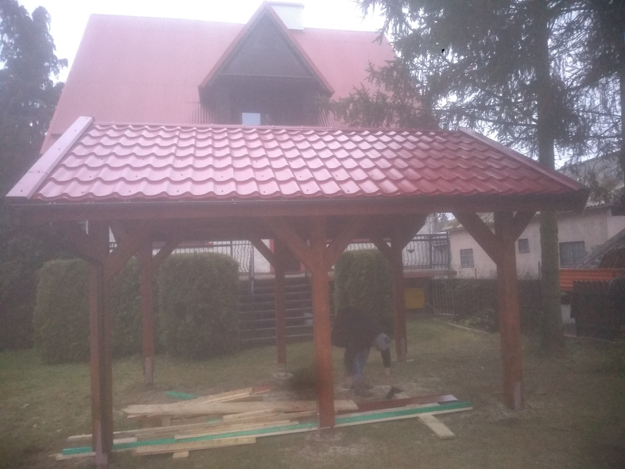 Montaż drewnianej altany z czerwoną dachówką na tle domu z czerwonym dachem, widoczne deski i pracownik w trakcie pracy.