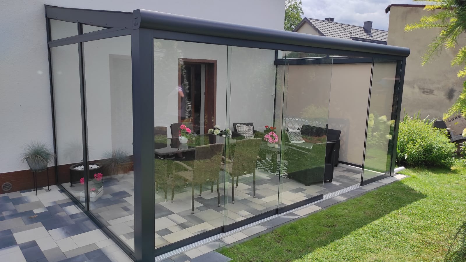Zadaszony taras z metalową konstrukcją i szklanymi ścianami, wyposażony w meble ogrodowe, kwiaty i dekoracje, z widokiem na trawnik i zabudowania mieszkalne w tle.
