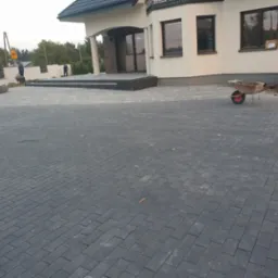 Ułożona, grafitowa kostka brukowa przed nowoczesnym domem z jasną elewacją, widoczna taczka i pracownicy w oddali.