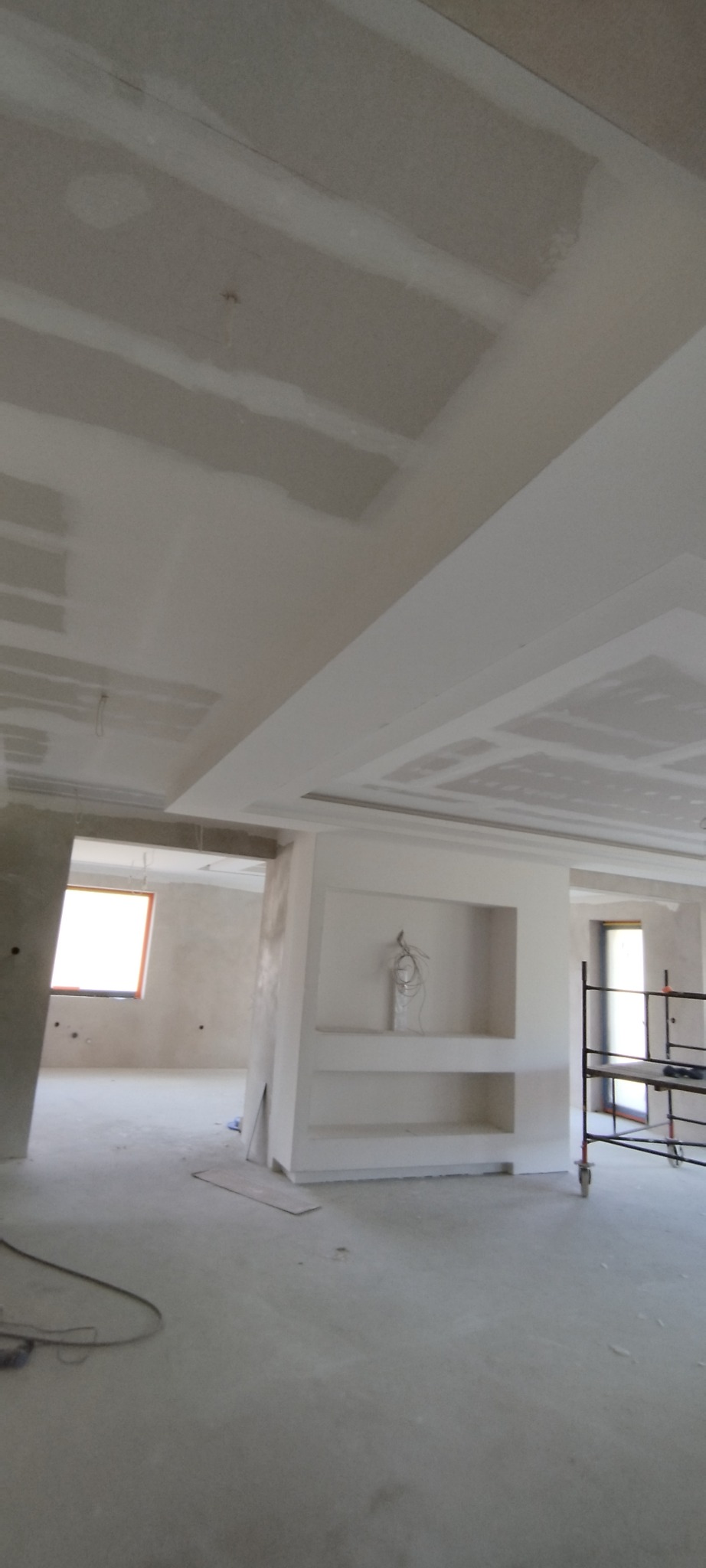 Wnętrze w trakcie prac wykończeniowych: sufit z płyt karton-gips, ściana działowa z wnękami, widoczne rusztowanie. Surowy stan, przygotowanie do malowania.