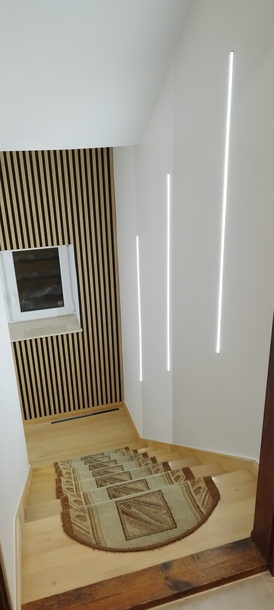 Nowoczesne schody z drewnianymi panelami ściennymi i oświetleniem LED w pionowych liniach. Dywaniki na schodach dodają przytulności.