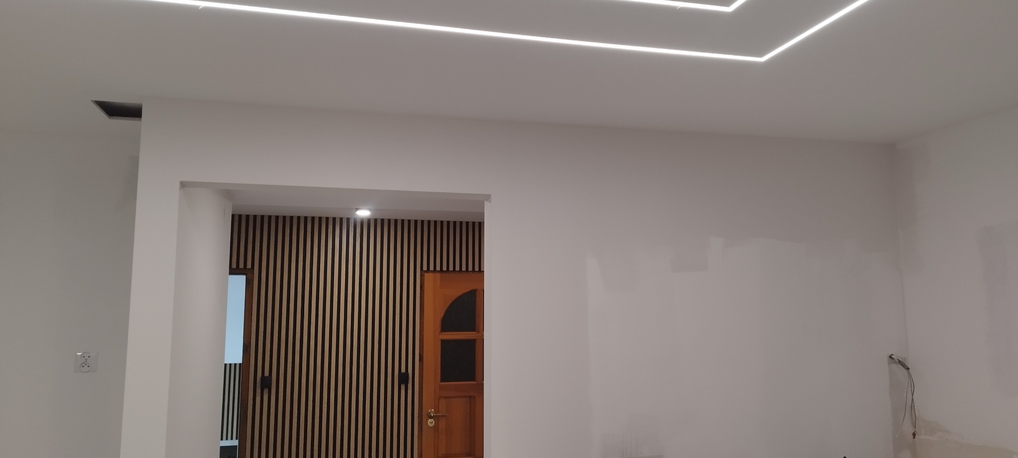 Białe wnętrze z nowoczesnym oświetleniem sufitowym LED w geometryczne wzory. Widoczna drewniana ściana i drzwi w tle. Minimalistyczny design.