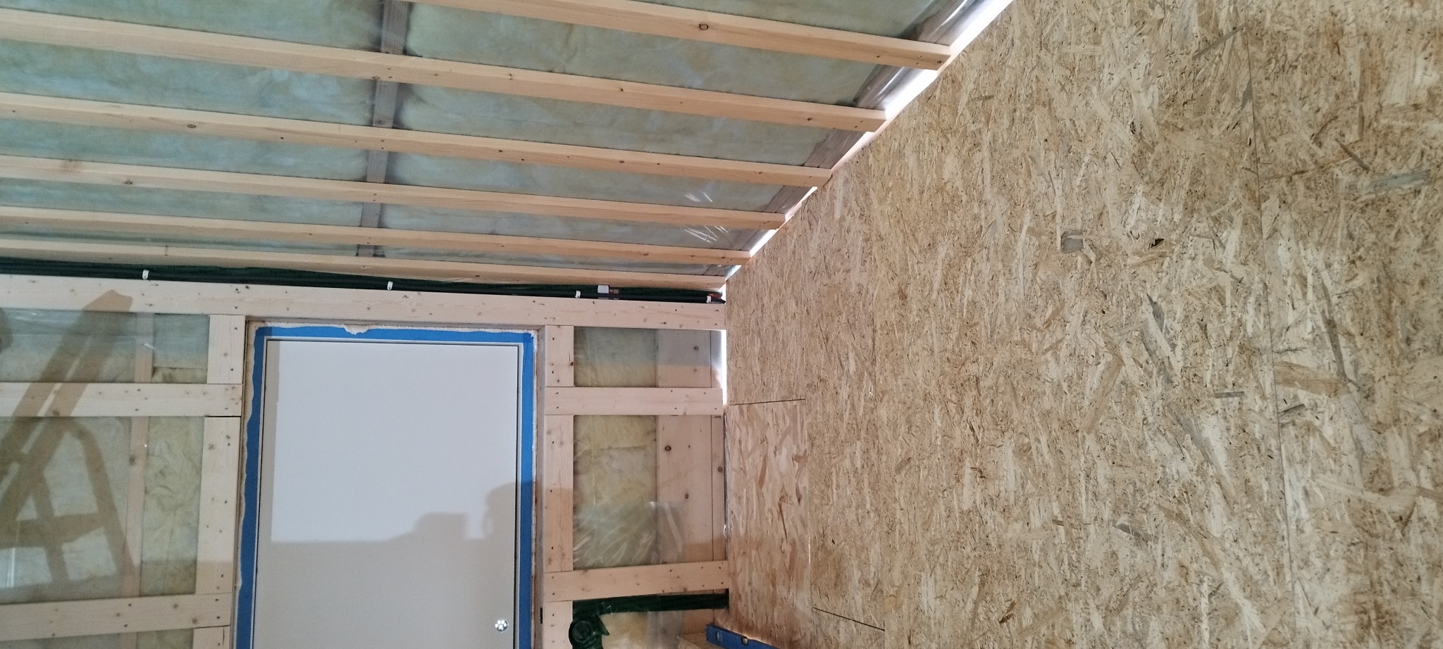 Wnętrze w trakcie budowy domu szkieletowego: widoczna konstrukcja drewniana, izolacja, płyta OSB na ścianie, fragment okna z niebieską taśmą zabezpieczającą.