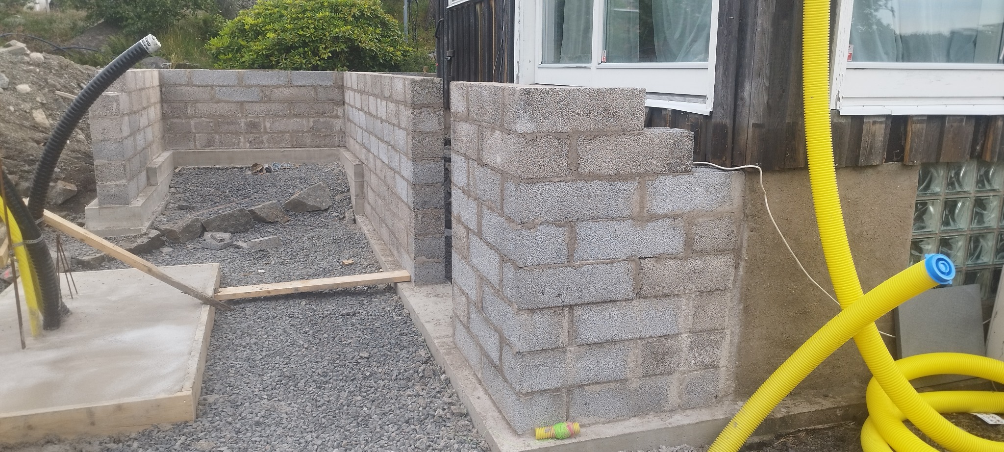 Wznoszenie ścian fundamentowych z bloczków betonowych. Widoczne żółte rury instalacyjne i podłoże z szarego żwiru. Częściowo ukończona konstrukcja.