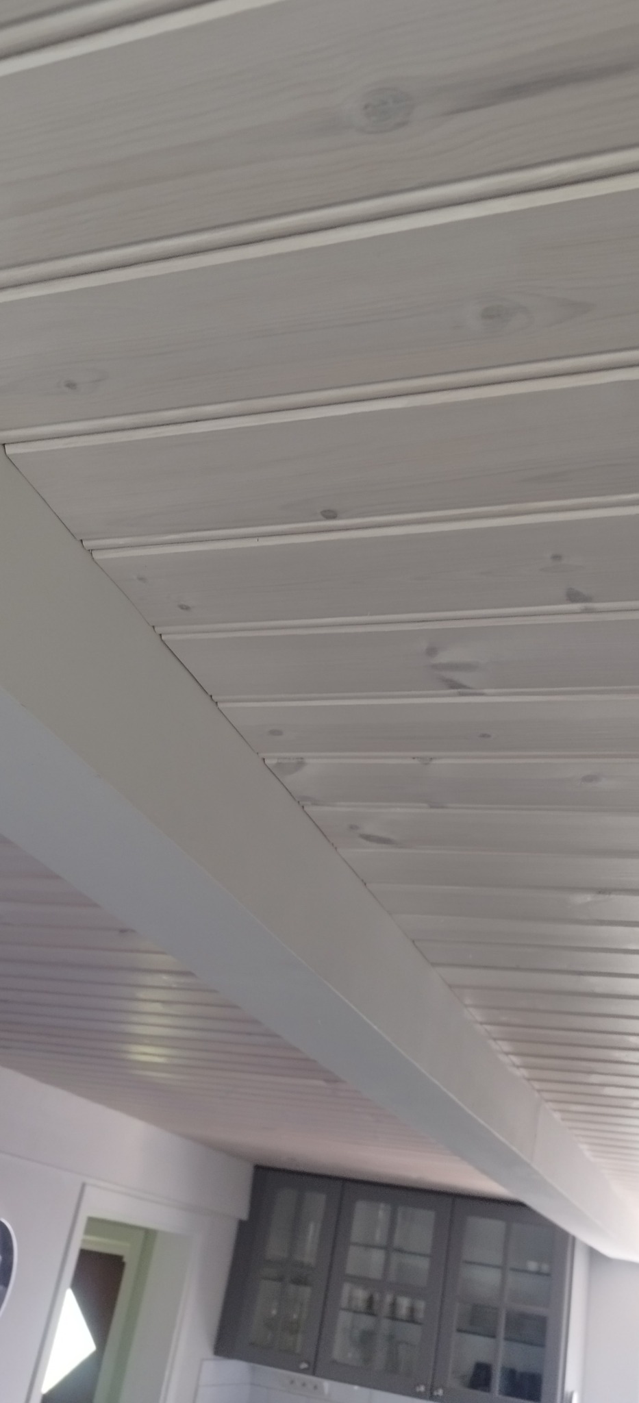 Jasnoszara, drewniana boazeria sufitowa z widocznymi sękami, zamontowana pod kątem. W tle fragment zabudowy kuchennej w szarym kolorze.