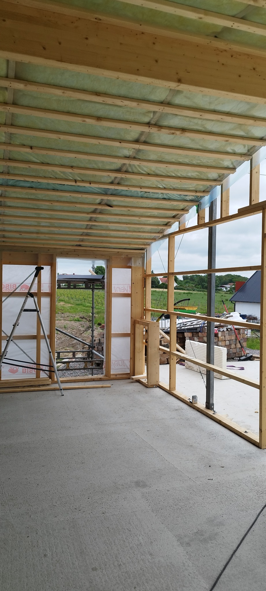 Wnętrze w budowie: surowy beton, drewniana konstrukcja szkieletowa z izolacją, drabina oparta o ścianę. Widok na zewnątrz przez otwory okienne, elementy budowlane na zewnątrz.