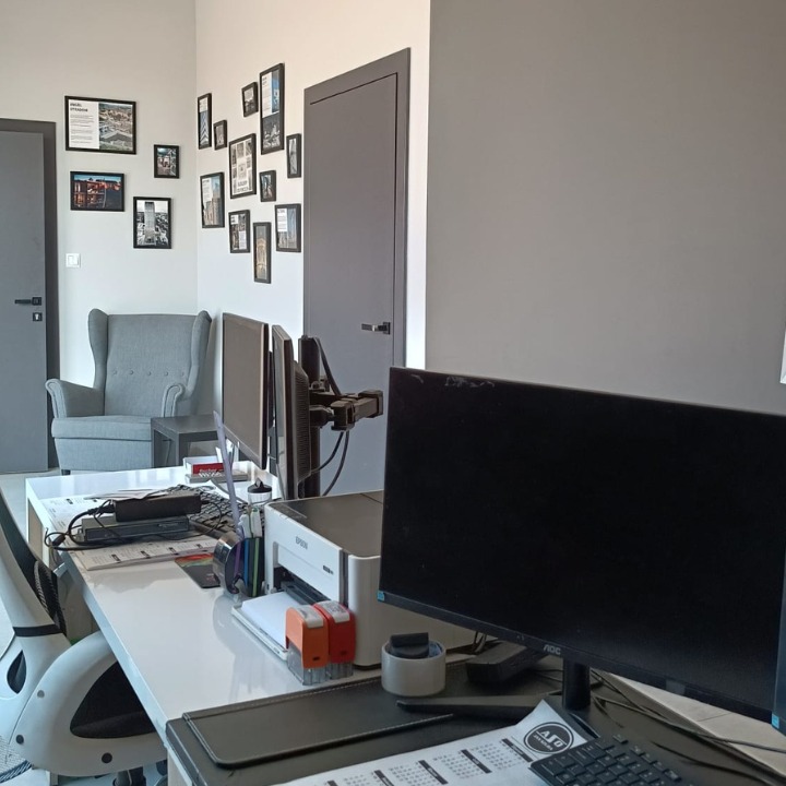 Biuro z białym biurkiem, dwoma monitorami, drukarką i ścianą z ramkami zdjęć w szarościach, widok zza biurka.