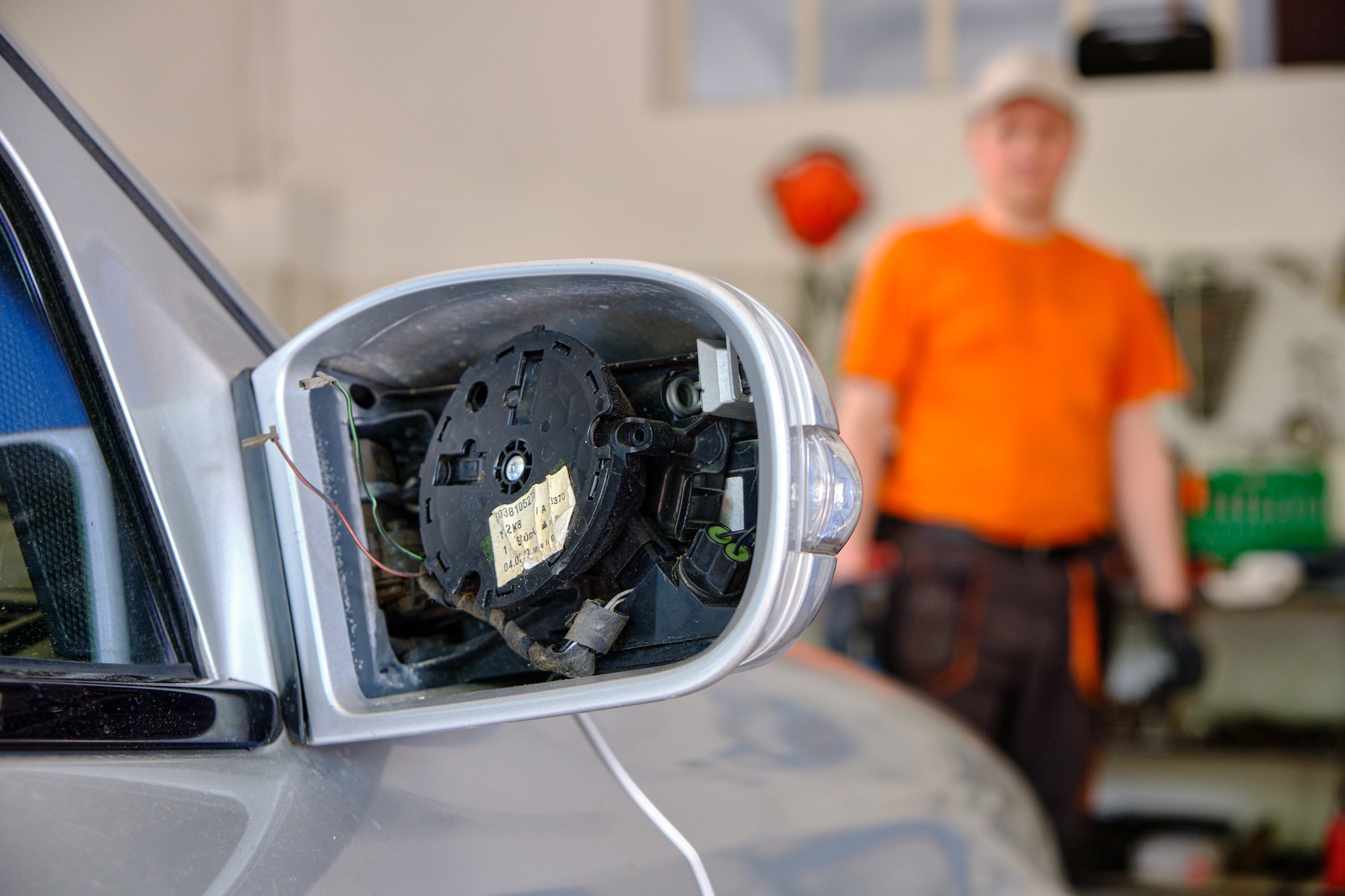 Uszkodzone lusterko boczne srebrnego samochodu w warsztacie, widoczne wewnętrzne mechanizmy i przewody; w tle zamazana postać mężczyzny w pomarańczowej koszulce i czarnych spodniach.