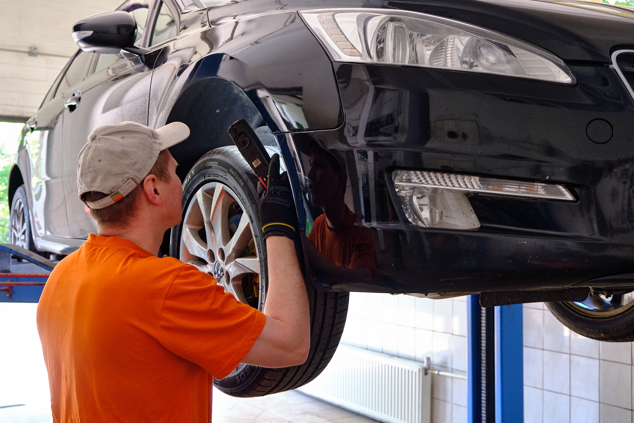 Czarny samochód na podnośniku w warsztacie w Gdańsku, mechanik w pomarańczowej koszulce i czarnych rękawicach sprawdza nadkole przy pomocy lampy inspekcyjnej.