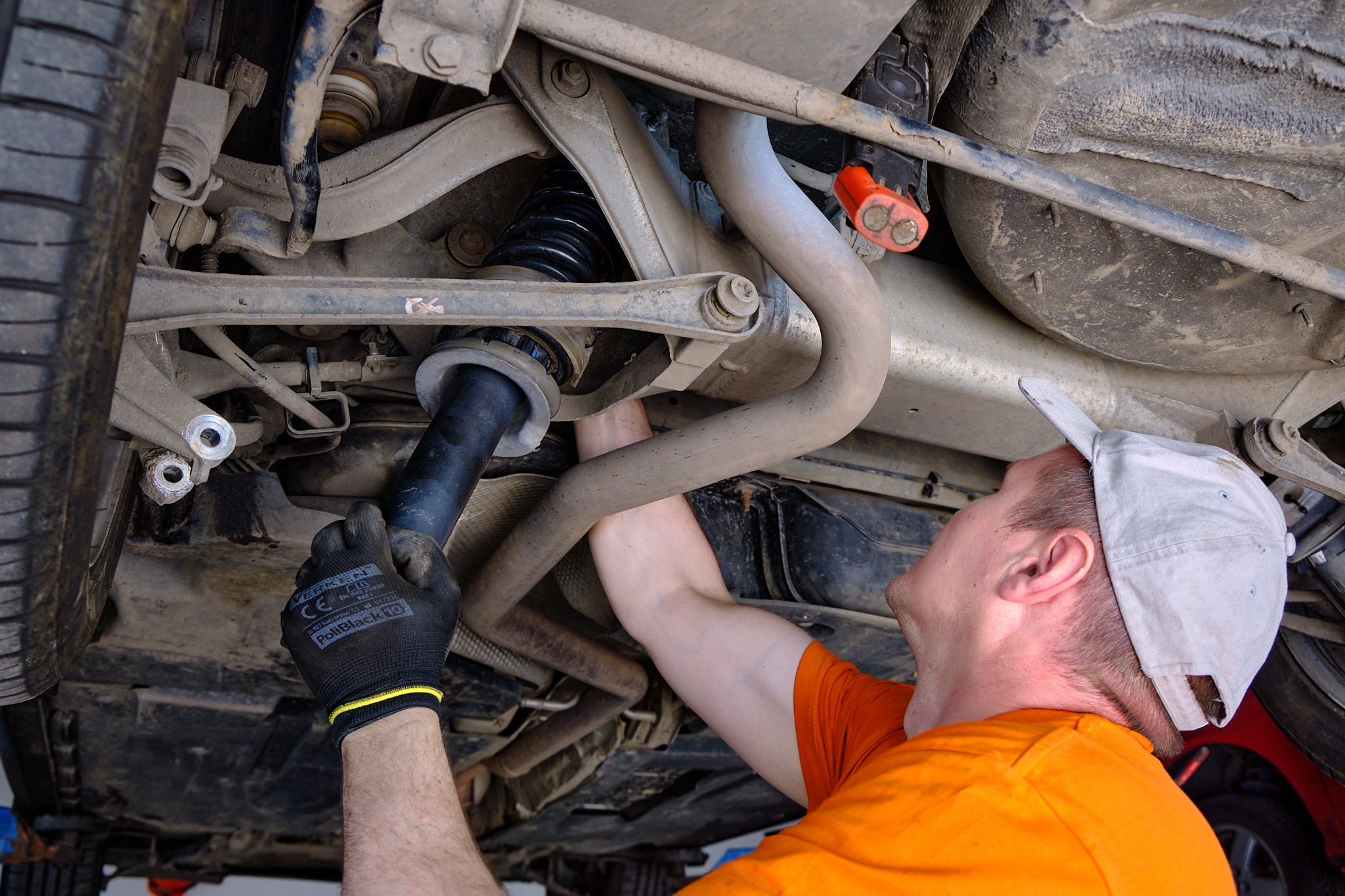 Człowiek w pomarańczowej koszulce i czarnych rękawicach roboczych montuje amortyzator w podwoziu samochodu na podnośniku, widoczne elementy zawieszenia i koło.