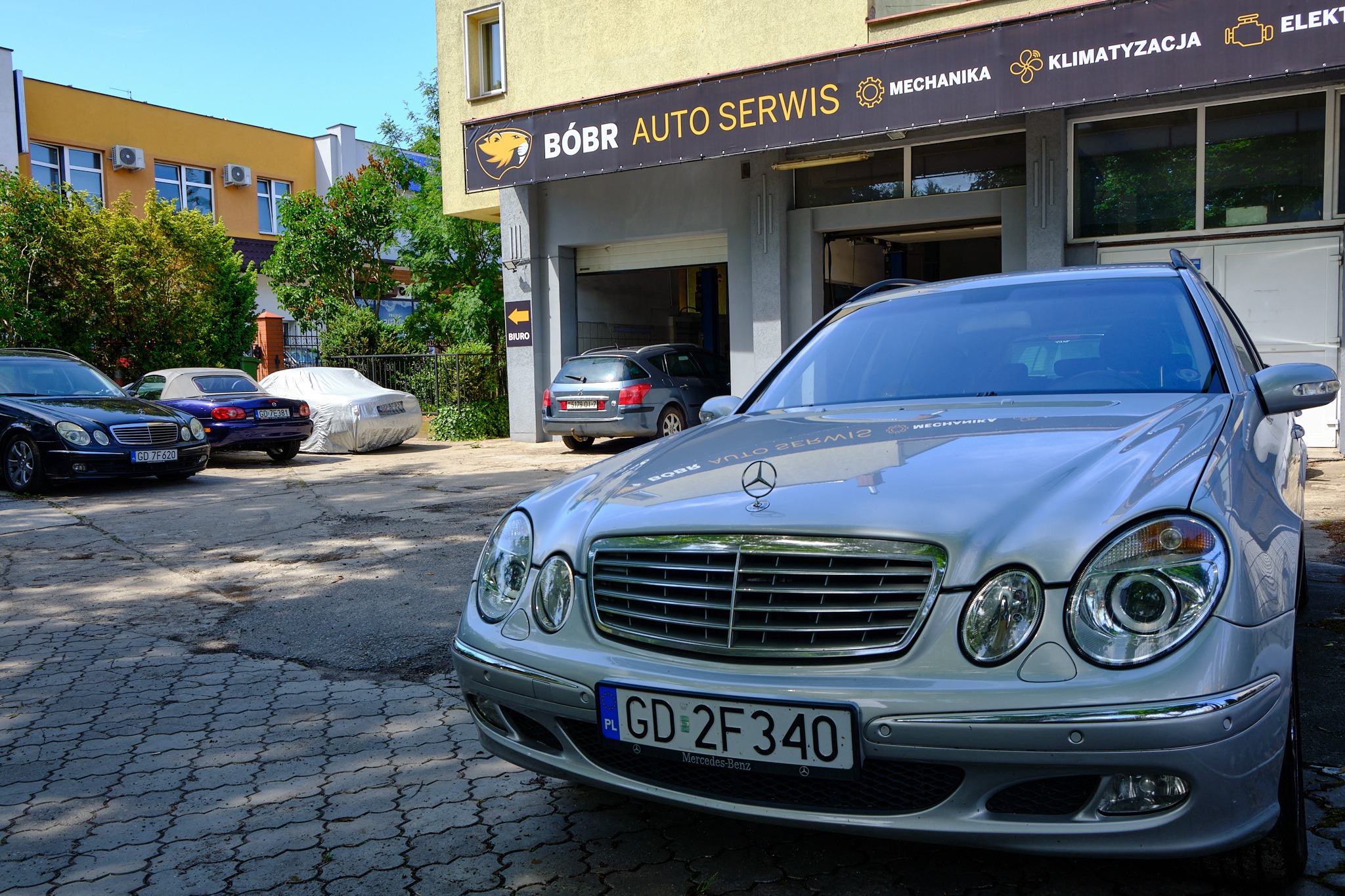 Srebrny Mercedes-Benz klasy E na pierwszym planie, zaparkowany przed warsztatem samochodowym 'Bóbr Auto Serwis' z widocznym szyldem oferującym mechanikę, klimatyzację i elektrykę; w tle inne...