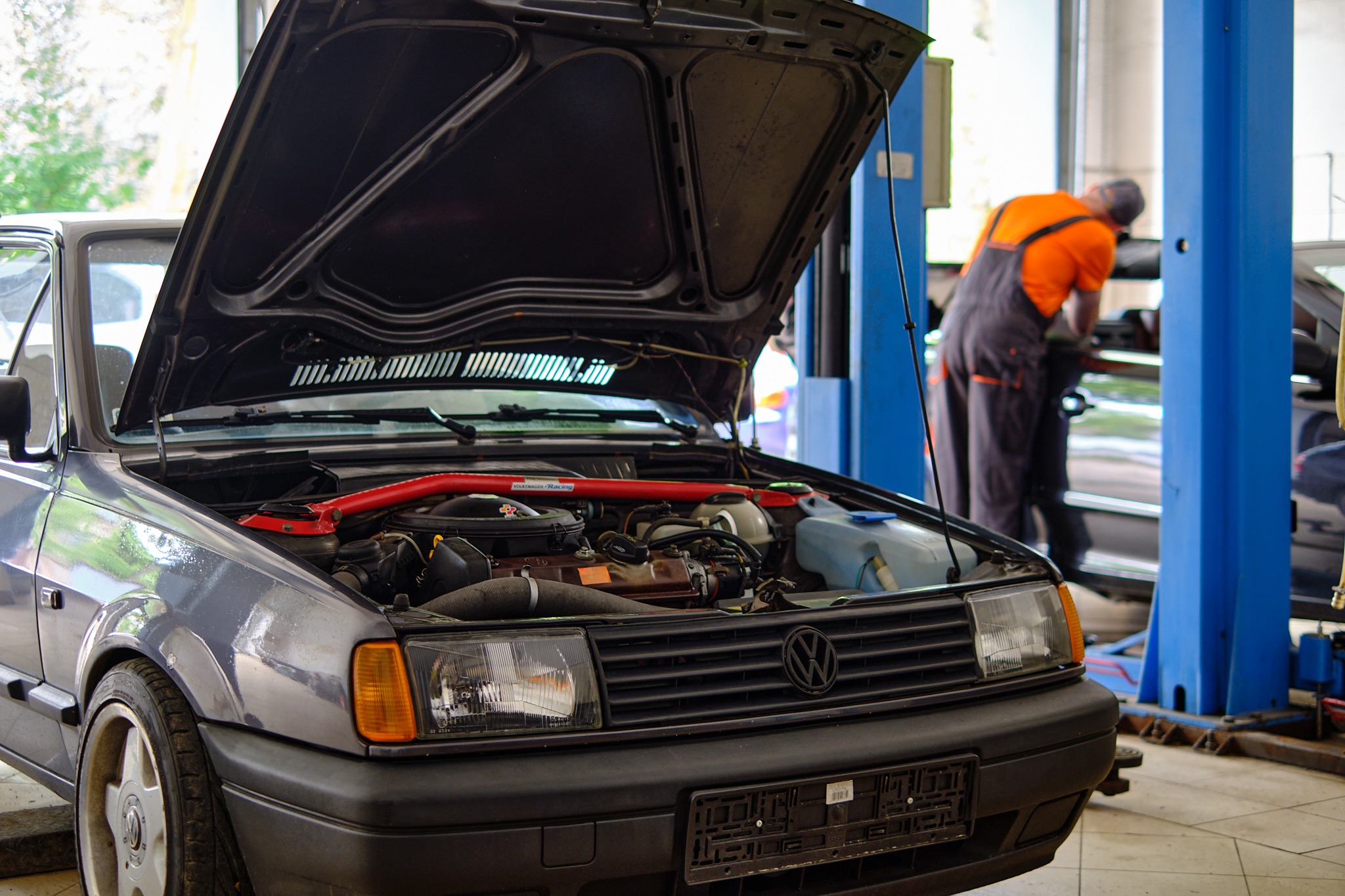 Szary Volkswagen Polo z otwartą maską w warsztacie; widoczny silnik z czerwoną rozpórką kielichów, mechanik w pomarańczowej koszulce pochylony nad innym autem w tle.