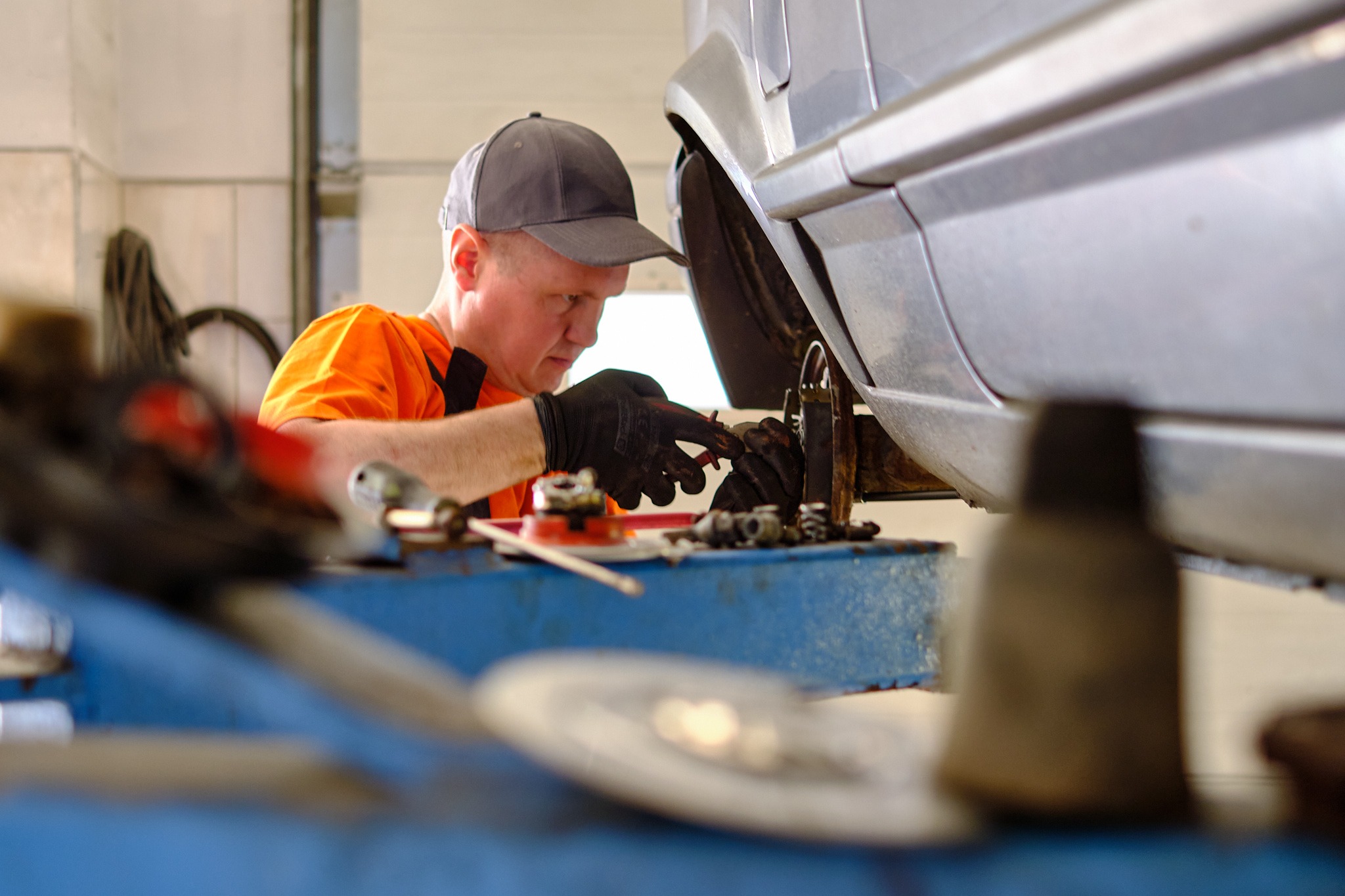 Mężczyzna w pomarańczowej koszulce i czarnych rękawicach pracuje przy podwoziu samochodu na podnośniku, skupiony na naprawie elementu zawieszenia, widoczne narzędzia i części samochodowe.