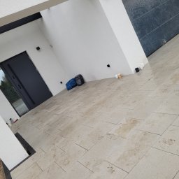 HUB-GARDEN-BRUK - Jasna kostka granitowa ułożona przed nowoczesnym budynkiem z ciemnymi akcentami. Widoczna brama wejściowa i fragment ściany z szarego kamienia.