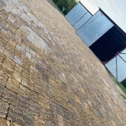 HUB-GARDEN-BRUK - Ukończona nawierzchnia z kostki granitowej w odcieniach beżu i szarości, prowadząca do blaszanych garaży. Nietypowy kąt ujęcia podkreśla fakturę kostki.