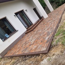 HUB-GARDEN-BRUK - Kaskadowa ścieżka z kostki brukowej w odcieniach brązu prowadzi do domu z oknami, na parapecie których stoją niebieskie kalosze dziecięce.