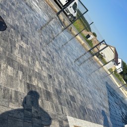 HUB-GARDEN-BRUK - Ułożona kostka brukowa na podjeździe z widocznym cieniem osoby. W tle biały bus i ogrodzenie z siatki.