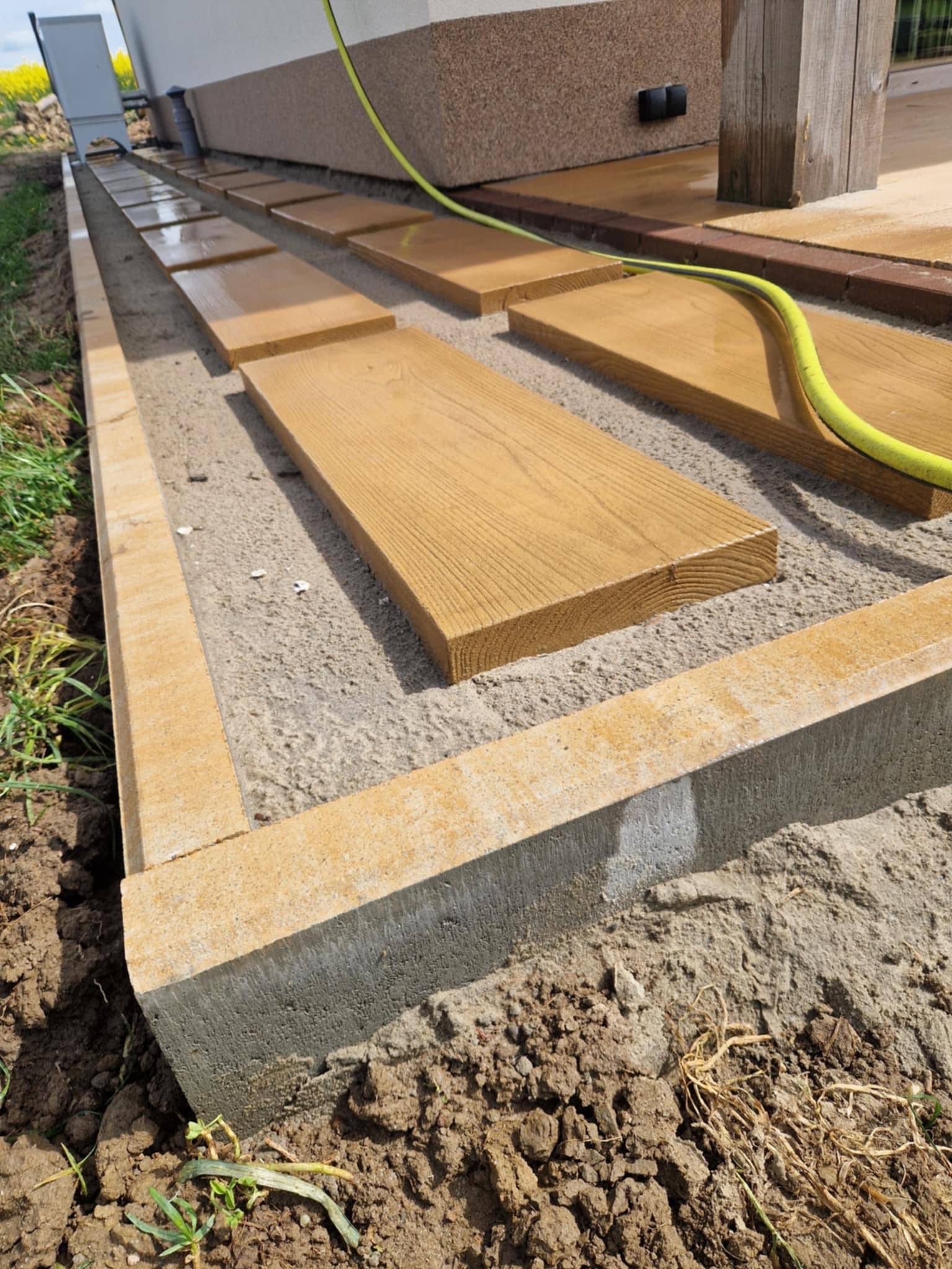 Przygotowanie podłoża pod ścieżkę z drewnianych desek. Obrzeża z betonowych kostek. Żółty wąż ogrodowy leży na deskach. Widok z góry, blisko ziemi.