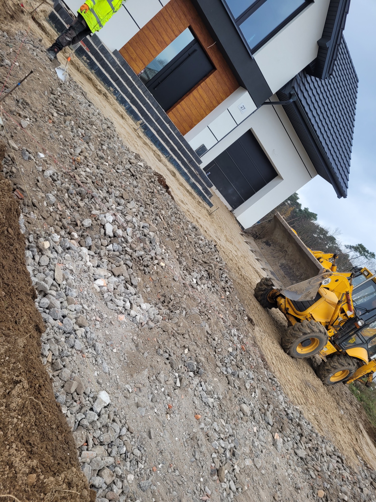 Żółta koparko-ładowarka na placu budowy przed nowoczesnym domem. Widoczne gruz, piasek i przygotowany teren pod utwardzenie. Pracownik w odblaskowej kamizelce.