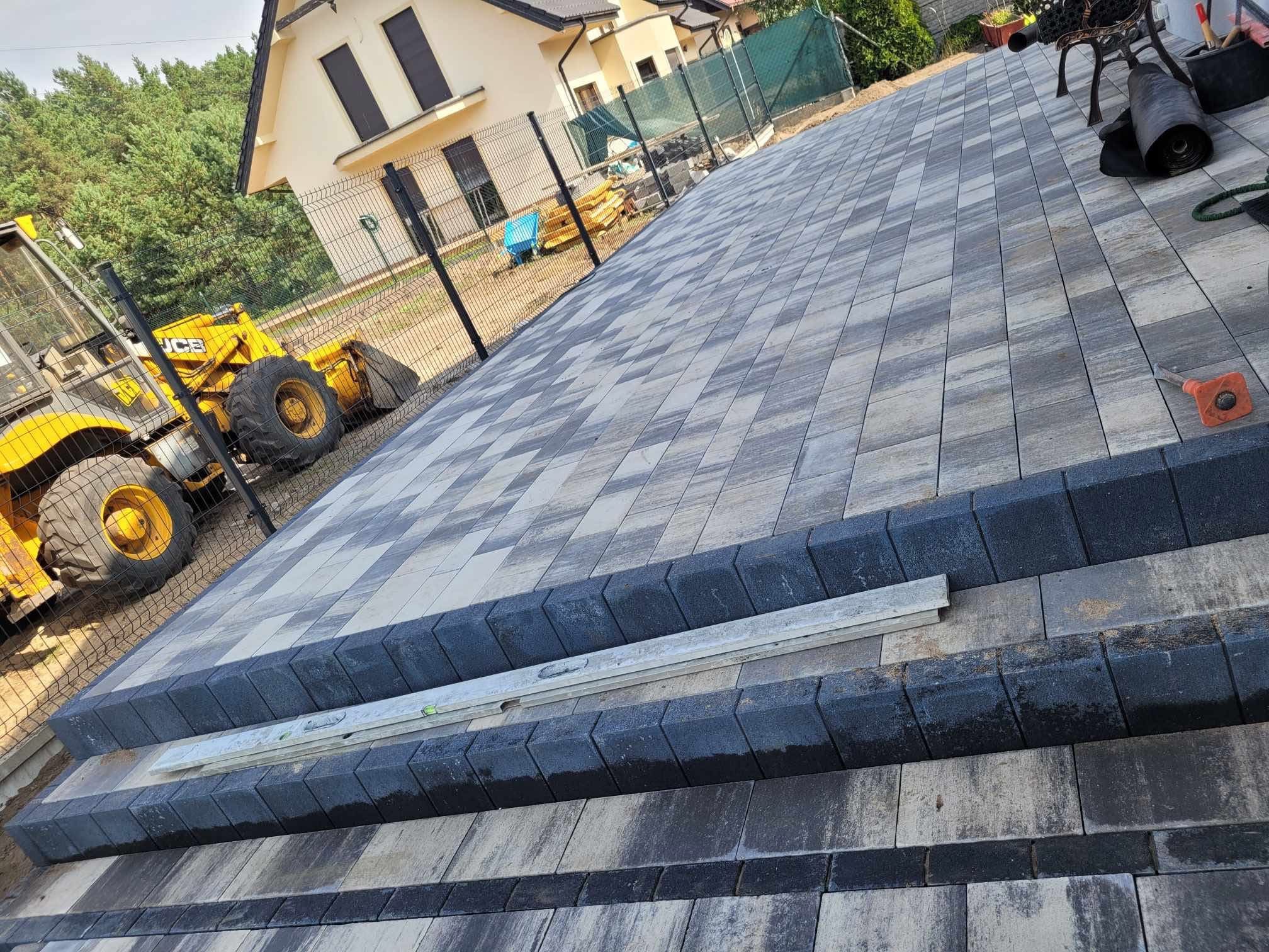 Ukończona nawierzchnia z kostki brukowej w szaro-białej kolorystyce, z obrzeżami w kolorze grafitowym. W tle widoczny budynek i maszyna budowlana.