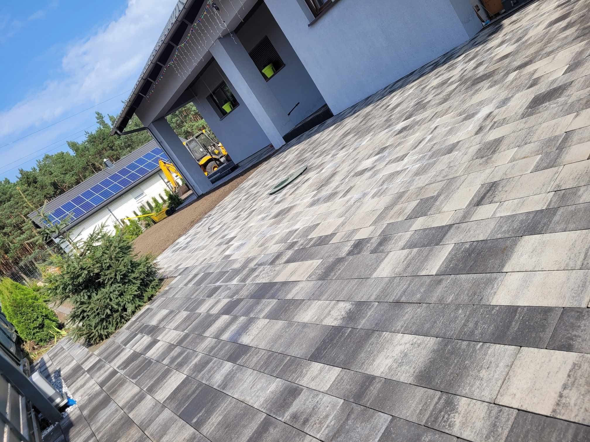 Ułożona kostka brukowa w odcieniach szarości, z widocznym budynkiem i sprzętem budowlanym w tle. Całość w jasnym, słonecznym oświetleniu.