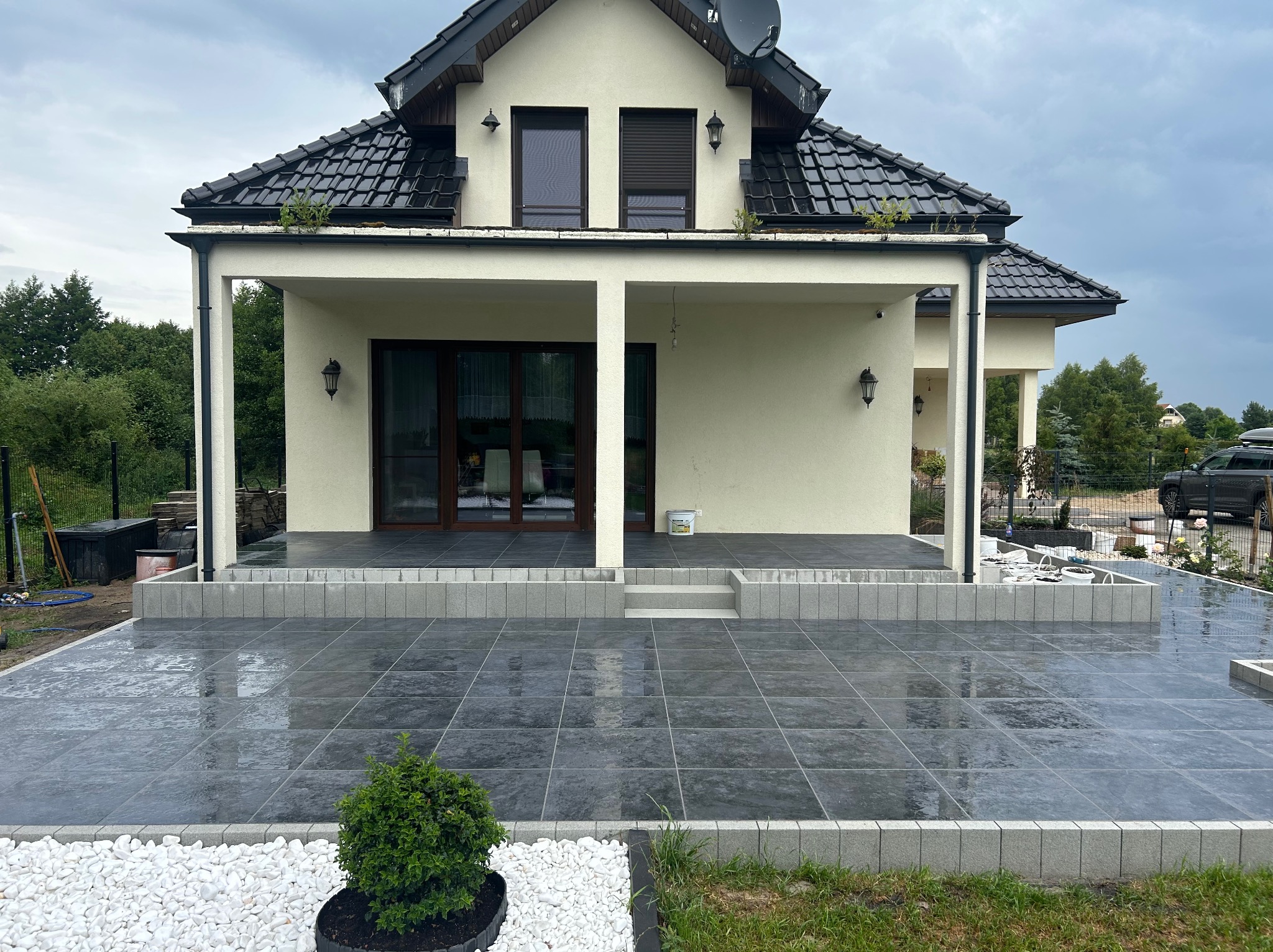 Nowoczesny taras z szarych płyt, obrzeża z kostki, białe kamienie dekoracyjne i zielony krzew na tle domu z kolumnami i czarnym dachem.