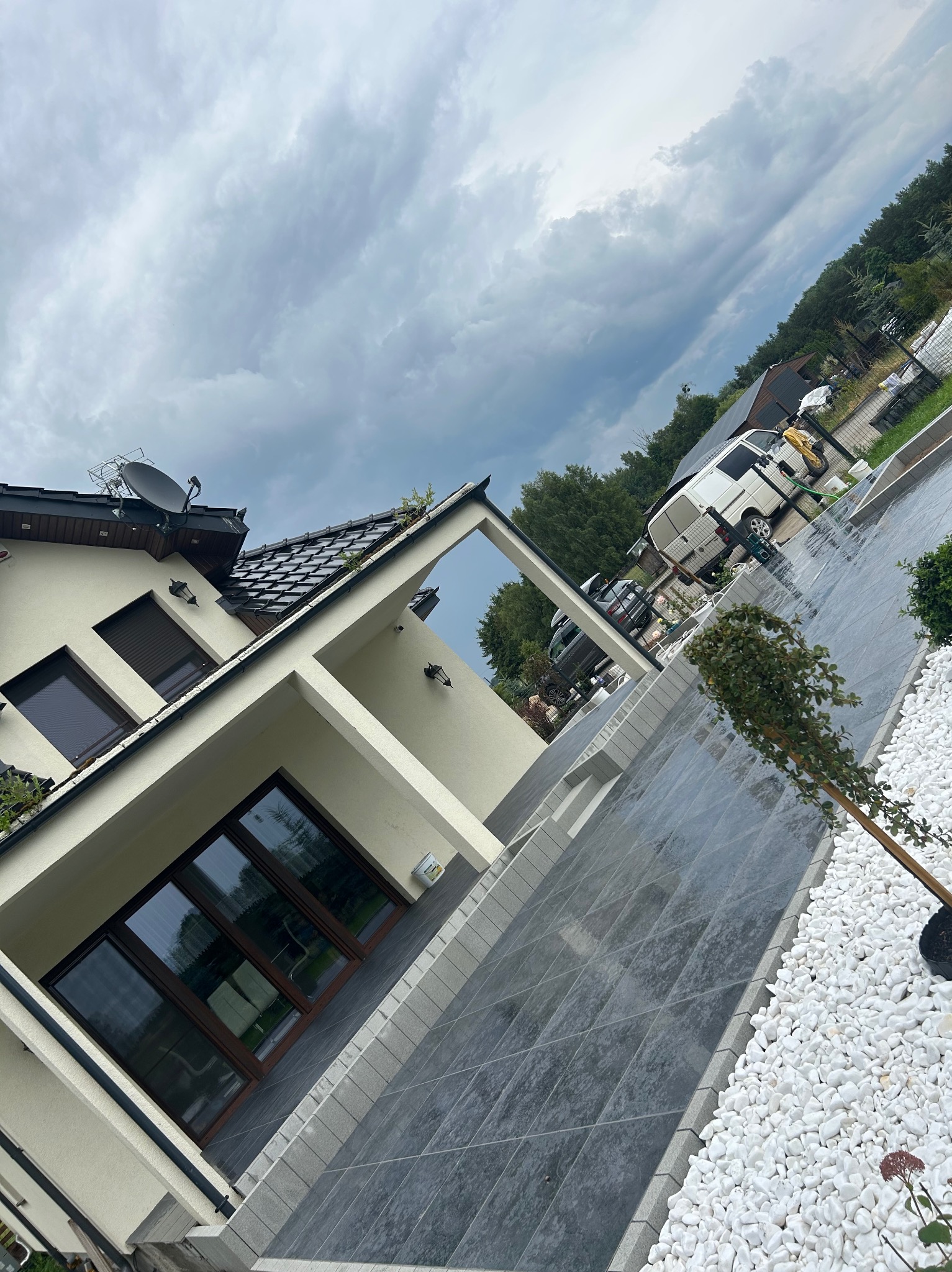 Nowoczesny podjazd z szarych płyt i białych kamieni ozdobnych, z widokiem na dom i zaparkowane samochody w tle, pochmurne niebo.