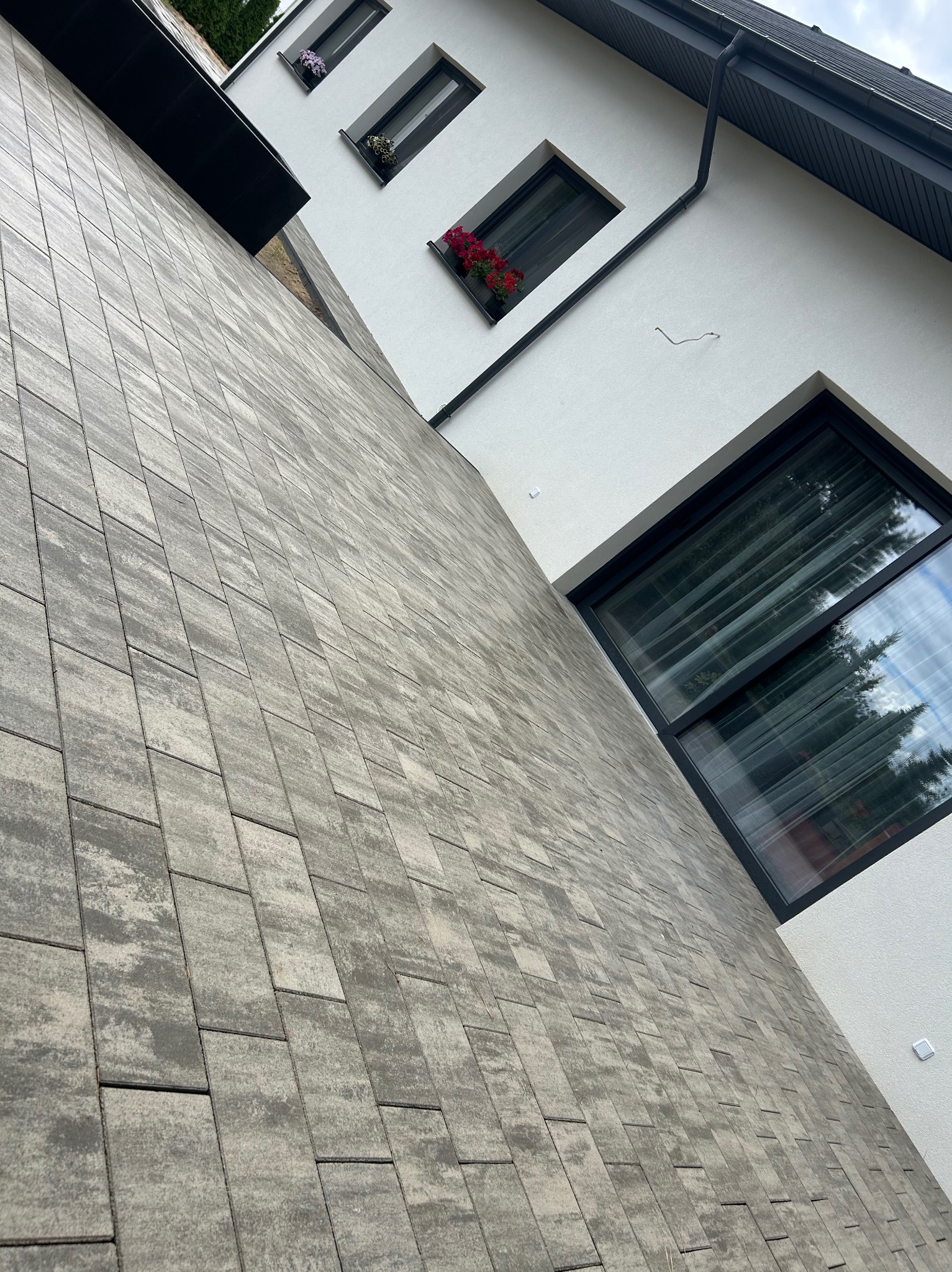 Ułożona kostka brukowa w odcieniach szarości przylegająca do białej ściany budynku z oknami. Widok ukośny, podkreślający wzór kostki i architekturę.