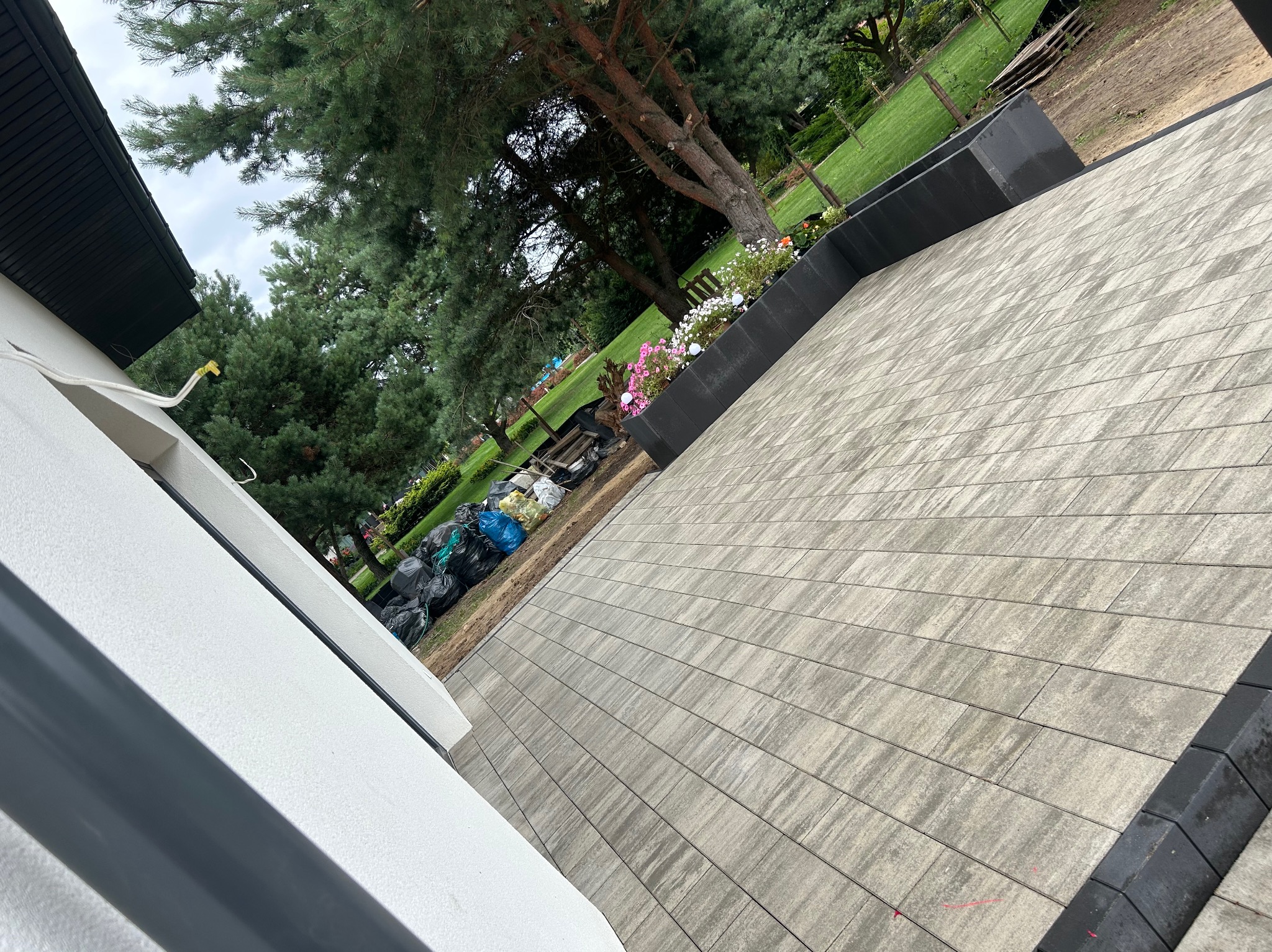 Ukończona nawierzchnia z kostki brukowej w odcieniach szarości, obrzeża z czarnej kostki. Widok z góry na ogród, drzewa i worki z ziemią w tle.