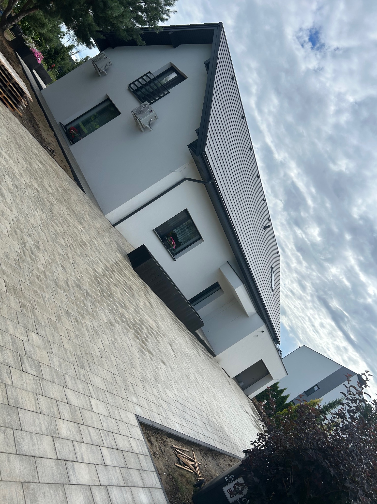 Nowoczesny dom z szarą kostką brukową na podjeździe, ujęcie z dołu, pochmurne niebo w tle. Widoczny fragment ogrodu i elewacja budynku.