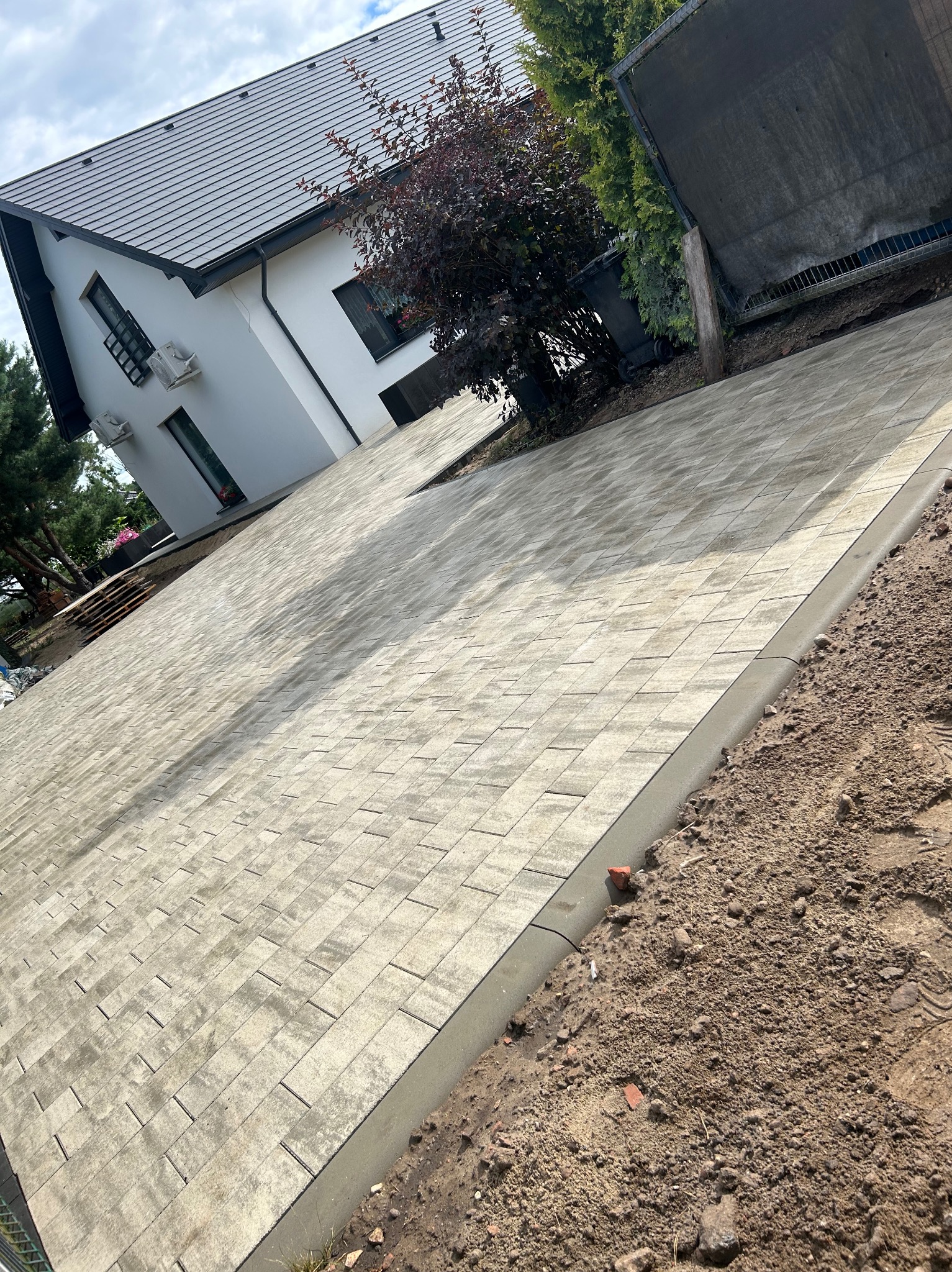 Nowoczesny podjazd z jasnoszarej kostki brukowej ułożonej wzorem prostokątnym, obrzeża betonowe, dom jednorodzinny w tle, częściowo widoczne drzewa i krzewy.