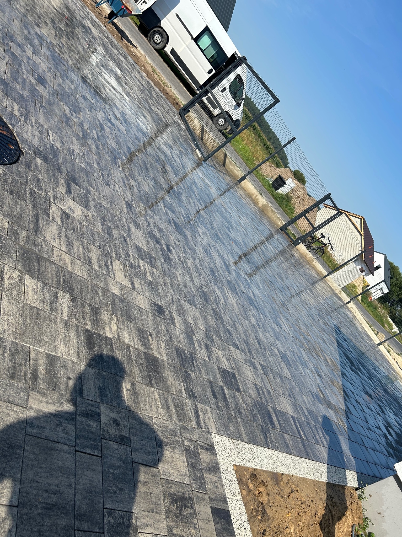 Ułożona kostka brukowa na podjeździe z widocznym cieniem osoby. W tle biały bus i ogrodzenie z siatki.