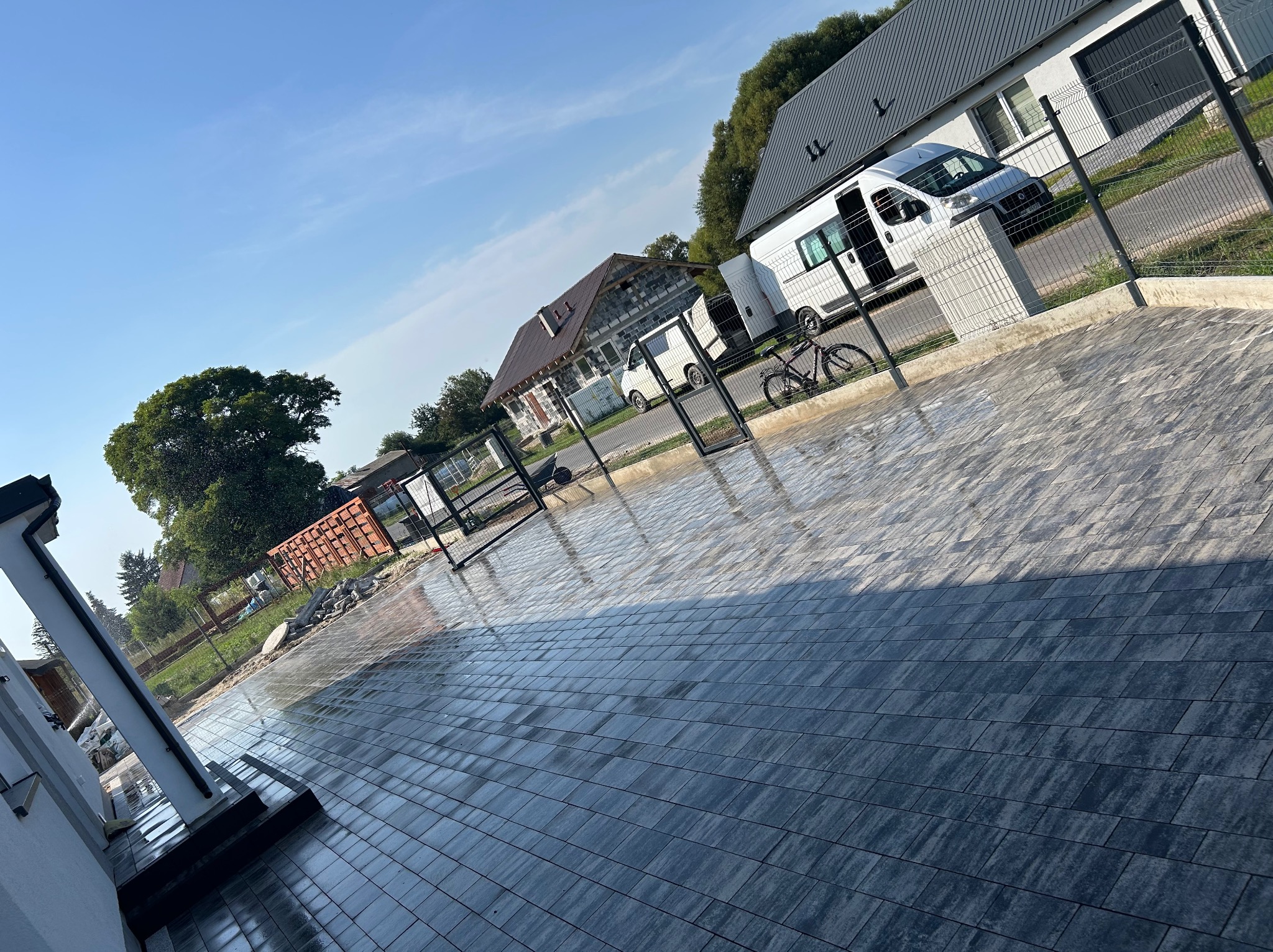 Ułożona, szara kostka brukowa na podjeździe domu jednorodzinnego, z widocznym ogrodzeniem panelowym i zaparkowanym busem. Świeżo wykonana nawierzchnia.