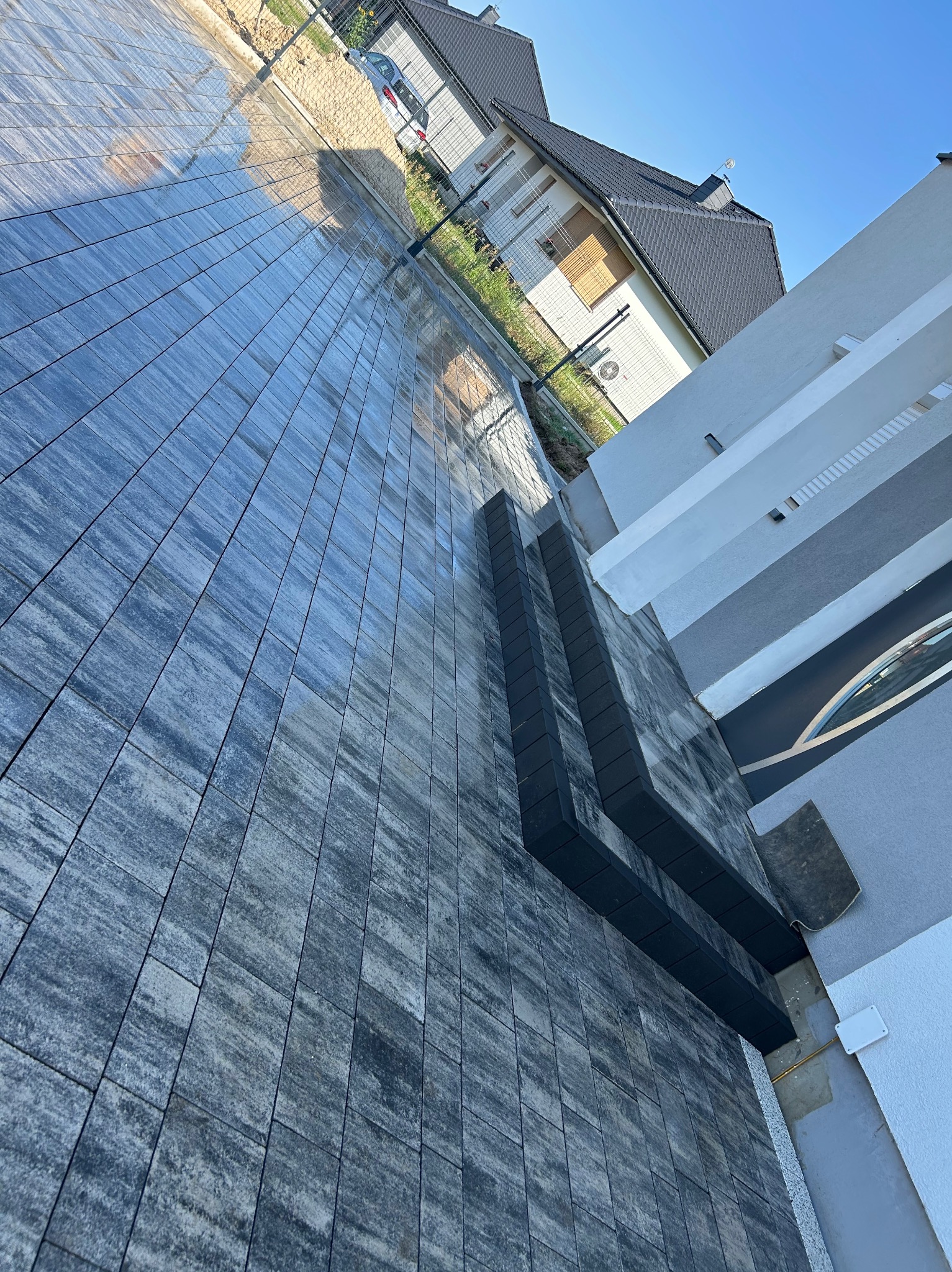 Ułożona, szara kostka brukowa z ciemnymi schodami na podjeździe domu w słoneczny dzień. Widok na budynek i samochód w tle.