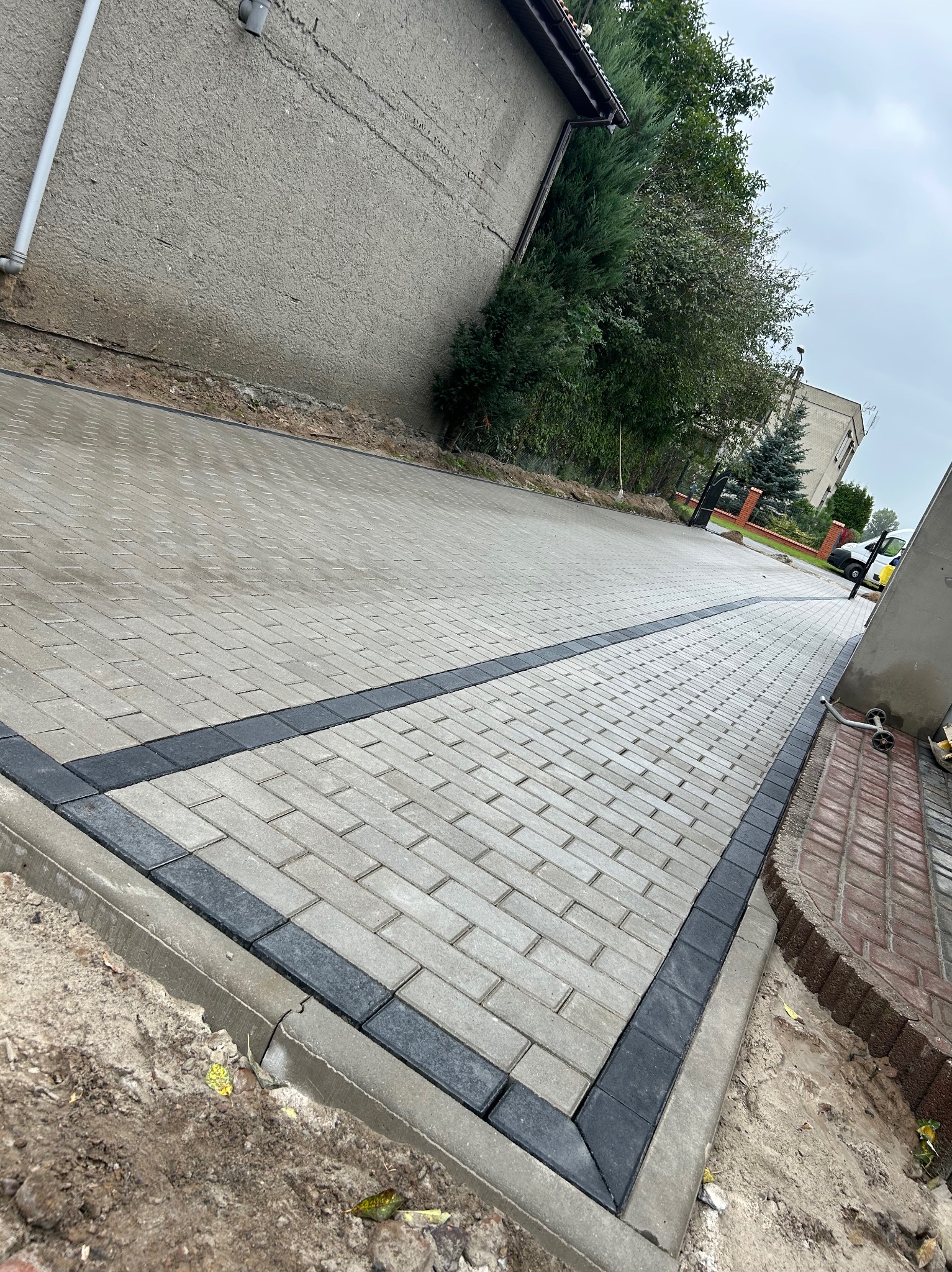 Ukończona nawierzchnia z jasnoszarej kostki brukowej, obramowana ciemnoszarą kostką. Widok na posesję z budynkiem i zielenią w tle, perspektywa z dołu.