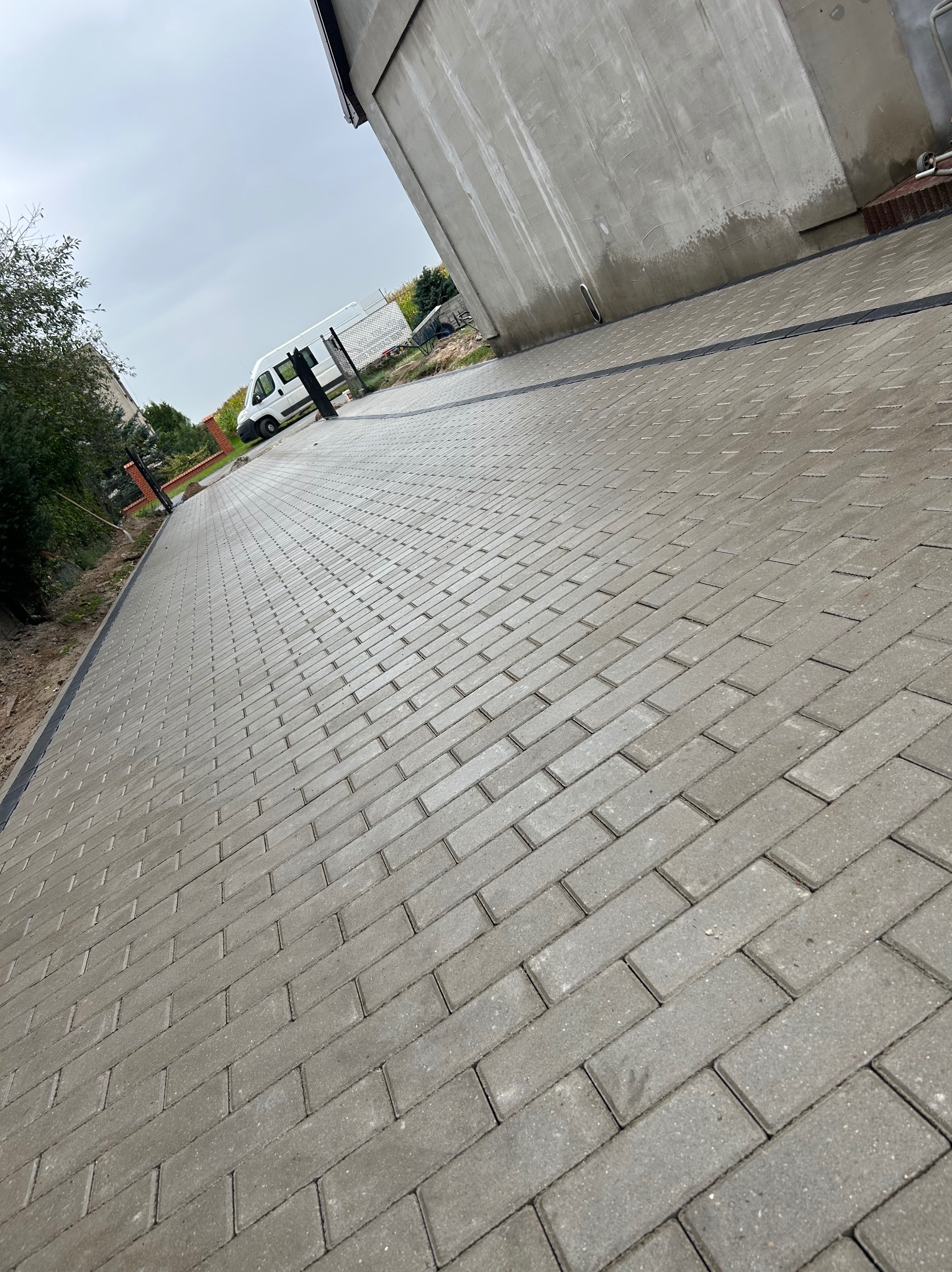 Ułożona, szara kostka brukowa w jodełkę, z widocznym drenażem liniowym wzdłuż budynku. W tle zaparkowany biały bus i zieleń.