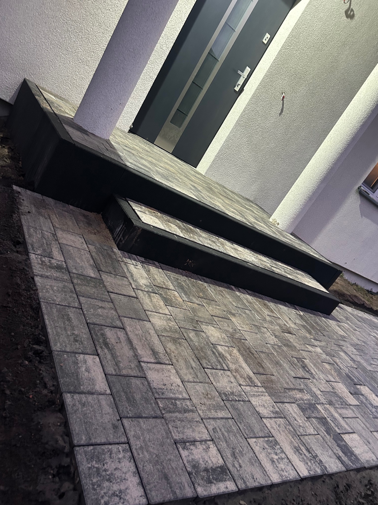 Ułożona kostka brukowa prowadząca do drzwi wejściowych z dwoma stopniami. Kostka w odcieniach szarości, czarna obramówka stopni, jasna elewacja budynku.