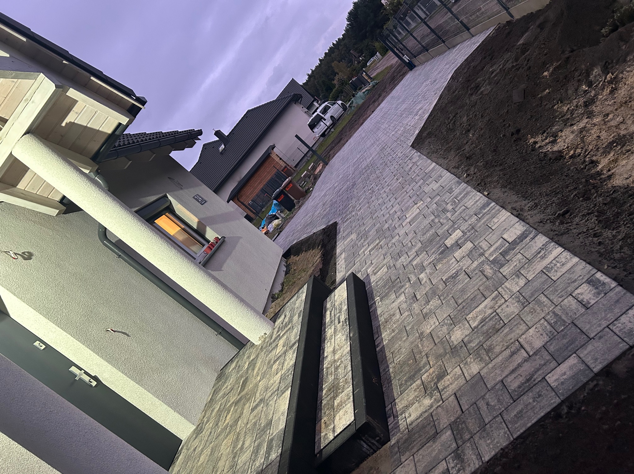 Ułożona, szara kostka brukowa prowadząca wzdłuż nowoczesnego domu, z widocznym fragmentem ogrodu i niebem w tle. Nietypowy kąt ujęcia podkreśla geometrię.