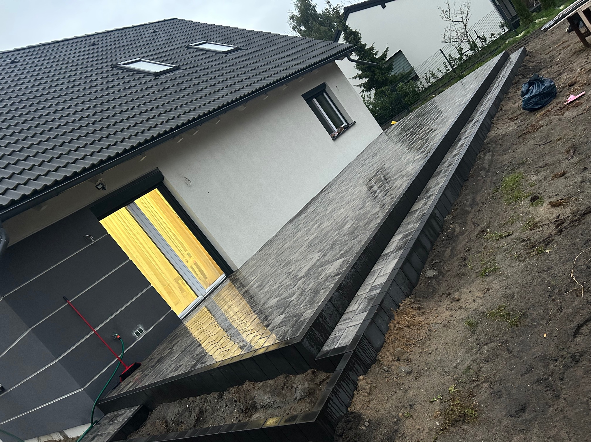 Ułożona kostka brukowa wzdłuż domu, z widocznymi obrzeżami. Kostka szara, mokra po deszczu. W tle dom z ciemną dachówką i fragment ogrodu.