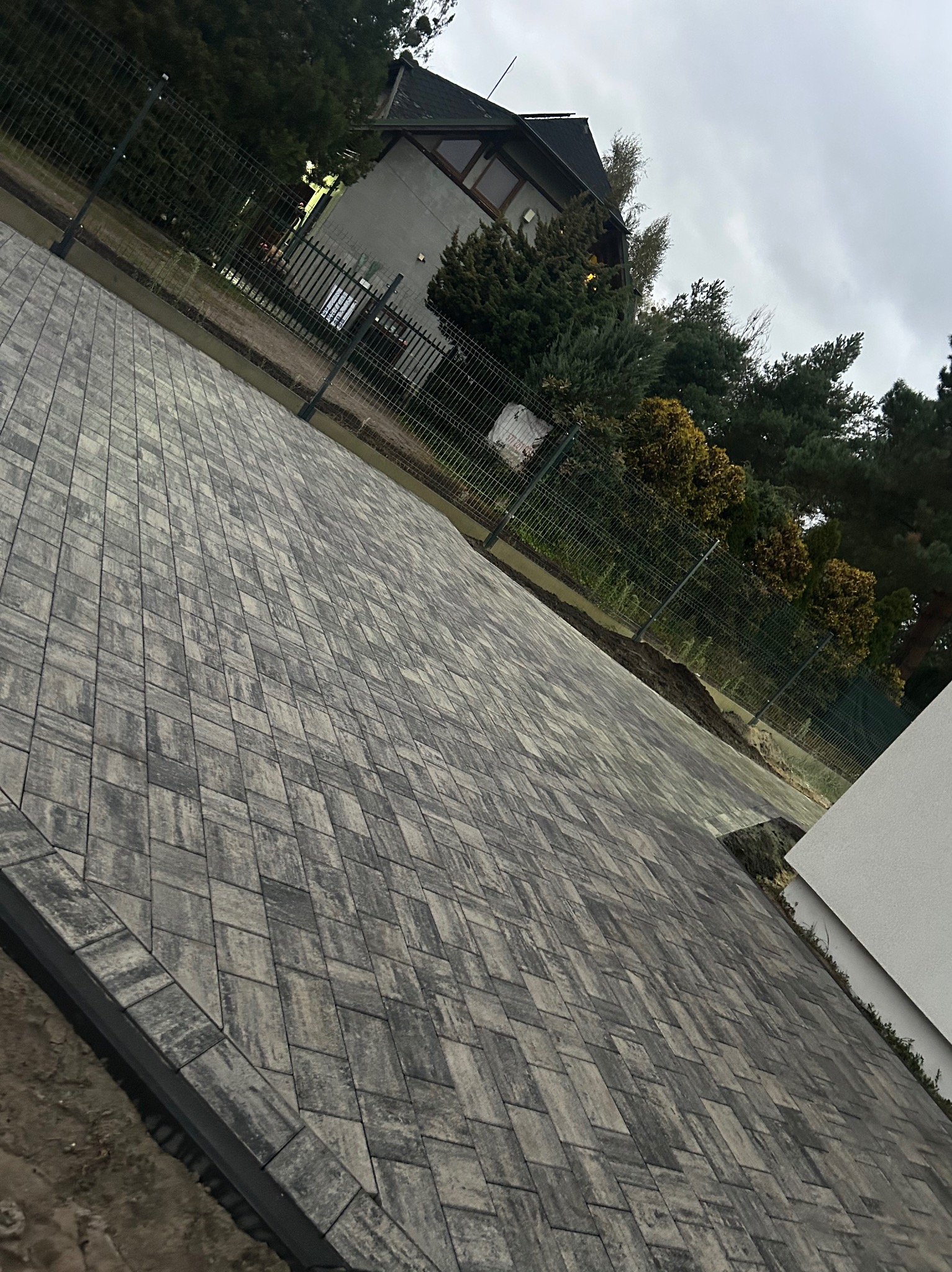 Ułożona kostka brukowa w jodełkę, obrzeża z palisady, widok na dom i ogrodzenie. Kostka w odcieniach szarości. Pochmurne niebo w tle.