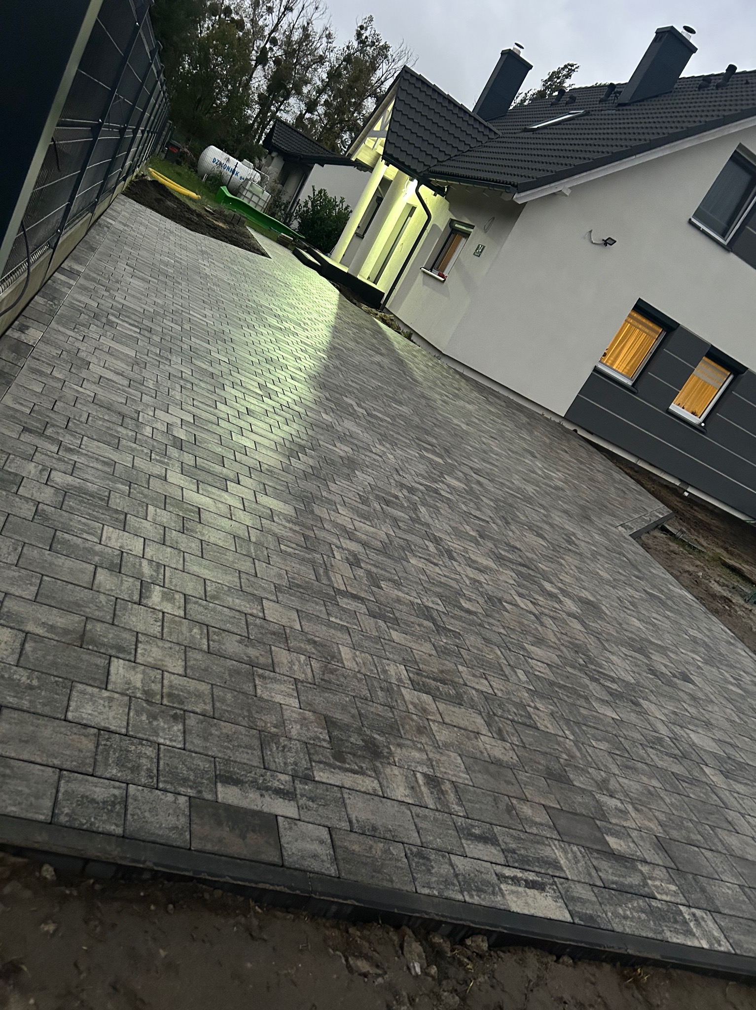 Ułożona kostka brukowa w szarościach prowadząca do nowoczesnego domu z oświetlonym wejściem, widok z niskiej perspektywy, pochmurne niebo.