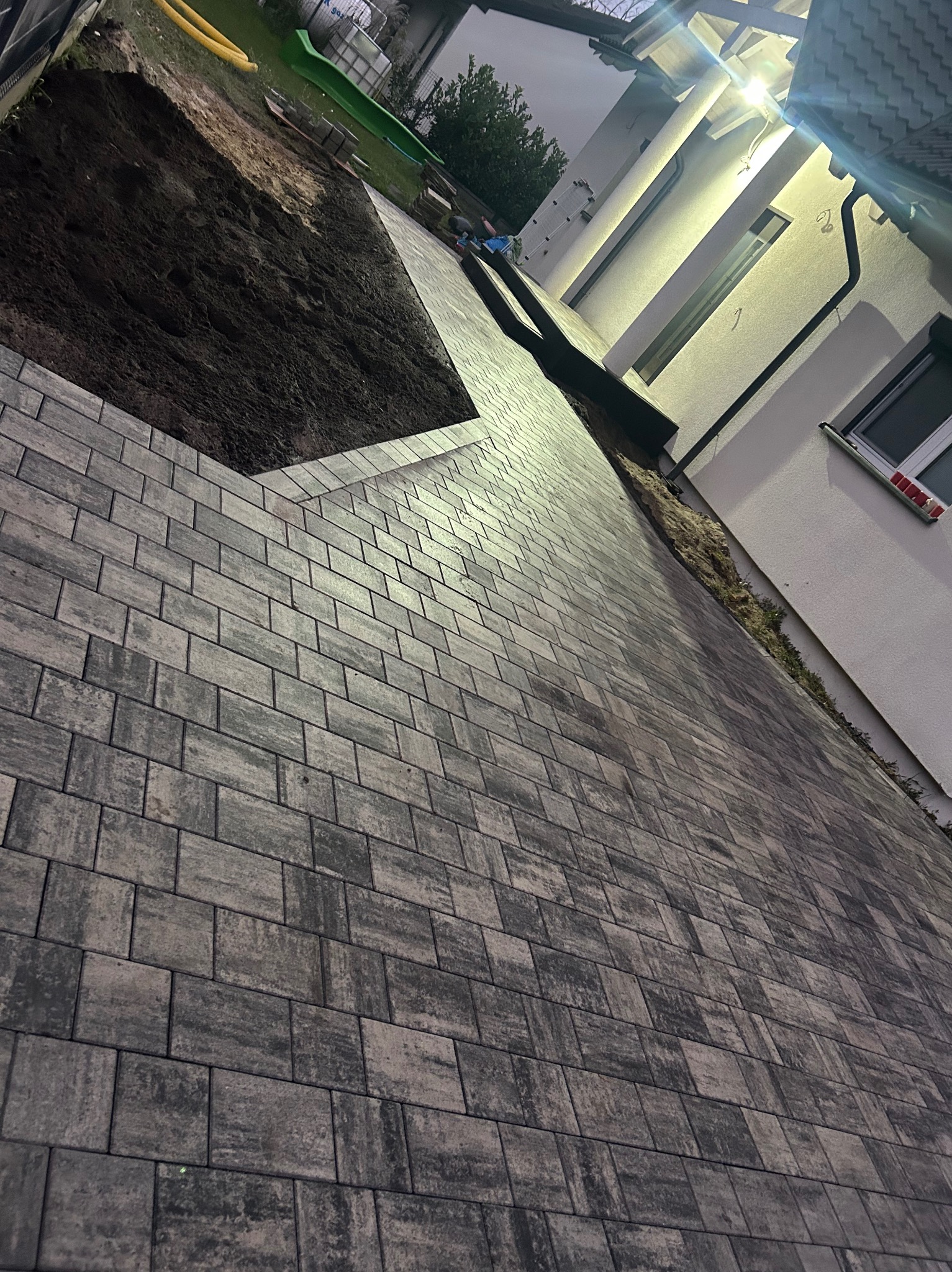 Ułożona, szara kostka brukowa na podjeździe posesji. Częściowo widoczna ziemia i budynek z oświetleniem. Niska perspektywa, ukazująca szczegóły faktury kostki.