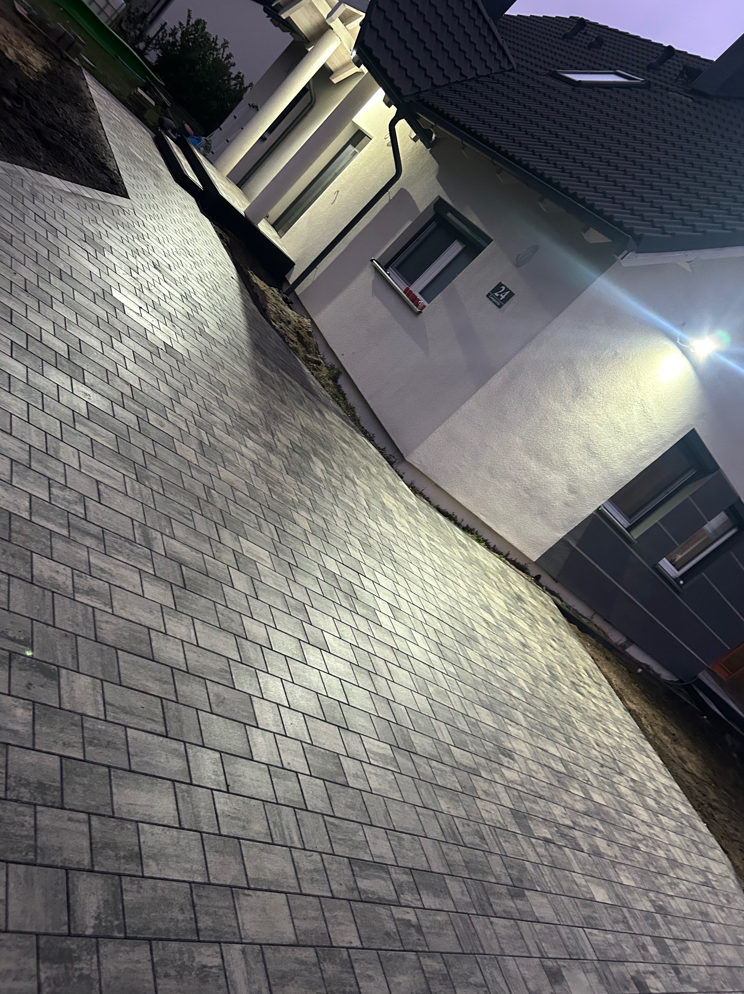 Nowo ułożona, szara kostka brukowa prowadząca do domu z białymi ścianami i ciemną dachówką, oświetlona światłem zewnętrznym.