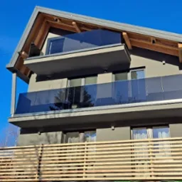 Nowoczesny dom z balkonami z szklanymi balustradami, widok z dołu na tle błękitnego nieba i częściowo drewnianego ogrodzenia.