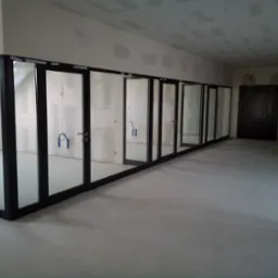 Długi korytarz w trakcie remontu z rzędem przeszklonych, czarnych drzwi przesuwnych, widoczne kable instalacji elektrycznej.